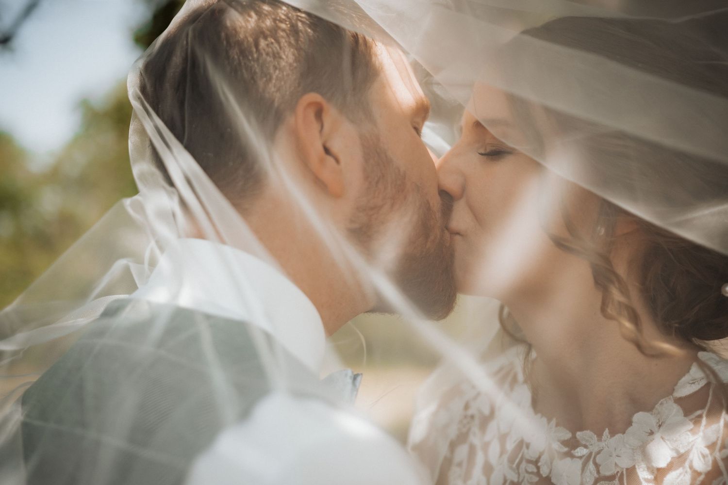 Bride and groom sharing a romantic kiss under a flowing white wedding veil outdoors on their wedding day.