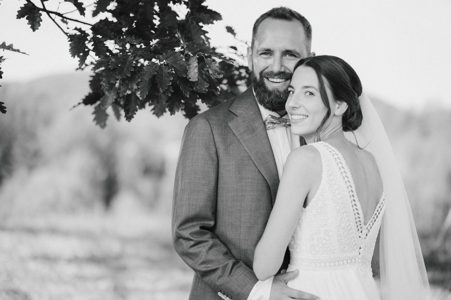 Black and white wedding photo of smiling bride and groom standing outdoors near a tree.
