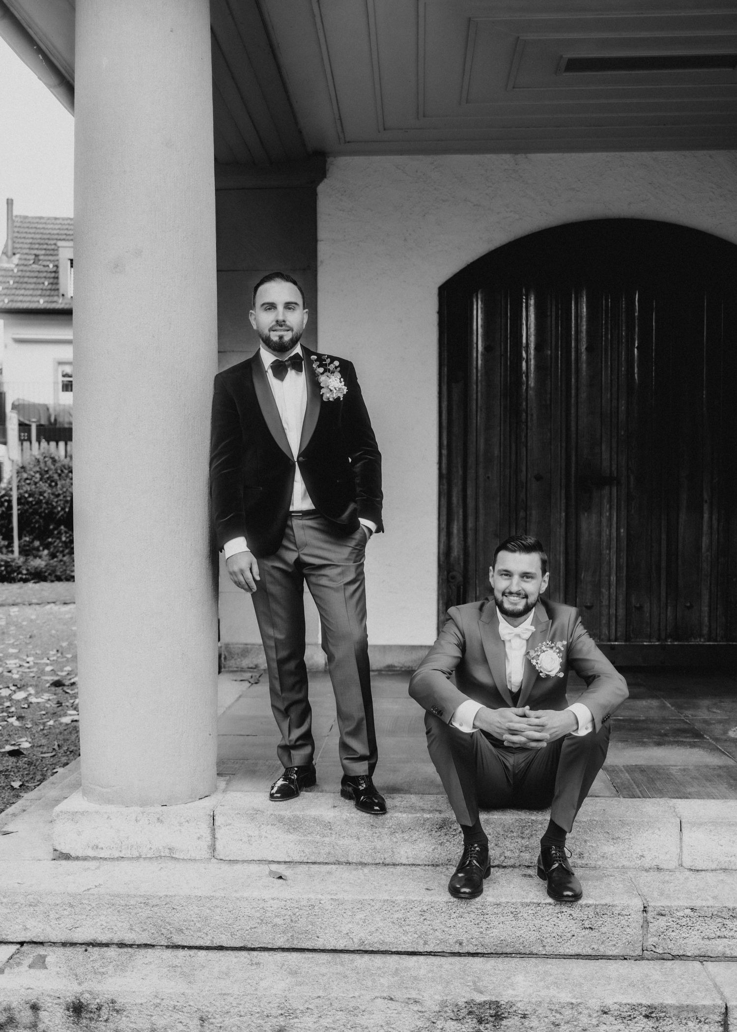 Black and white portrait of two men in formal attire posing on steps of a building with columns and arched doorway.