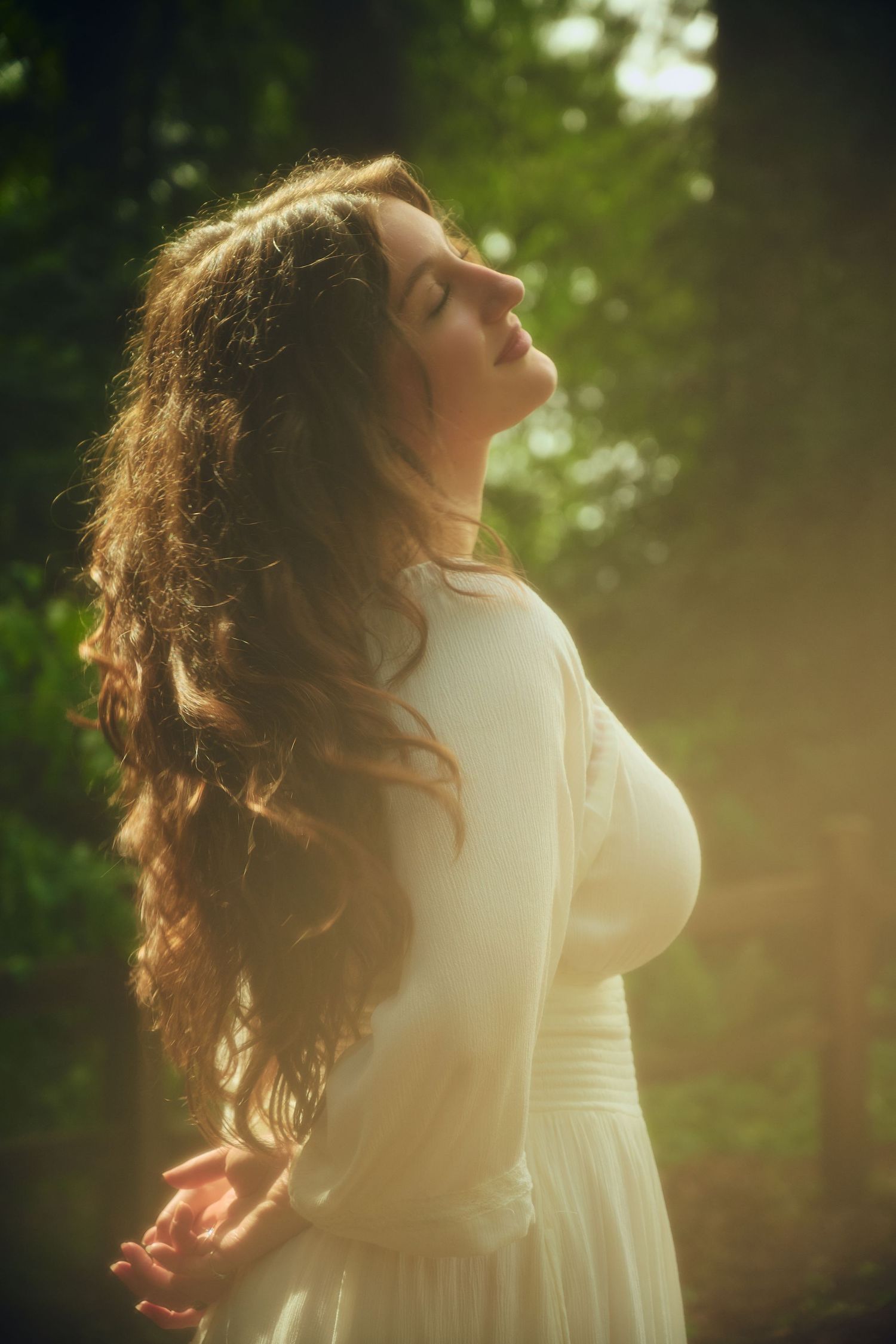 Woman in white dress with long curly hair, eyes closed, basking in sunlight in a lush green forest.