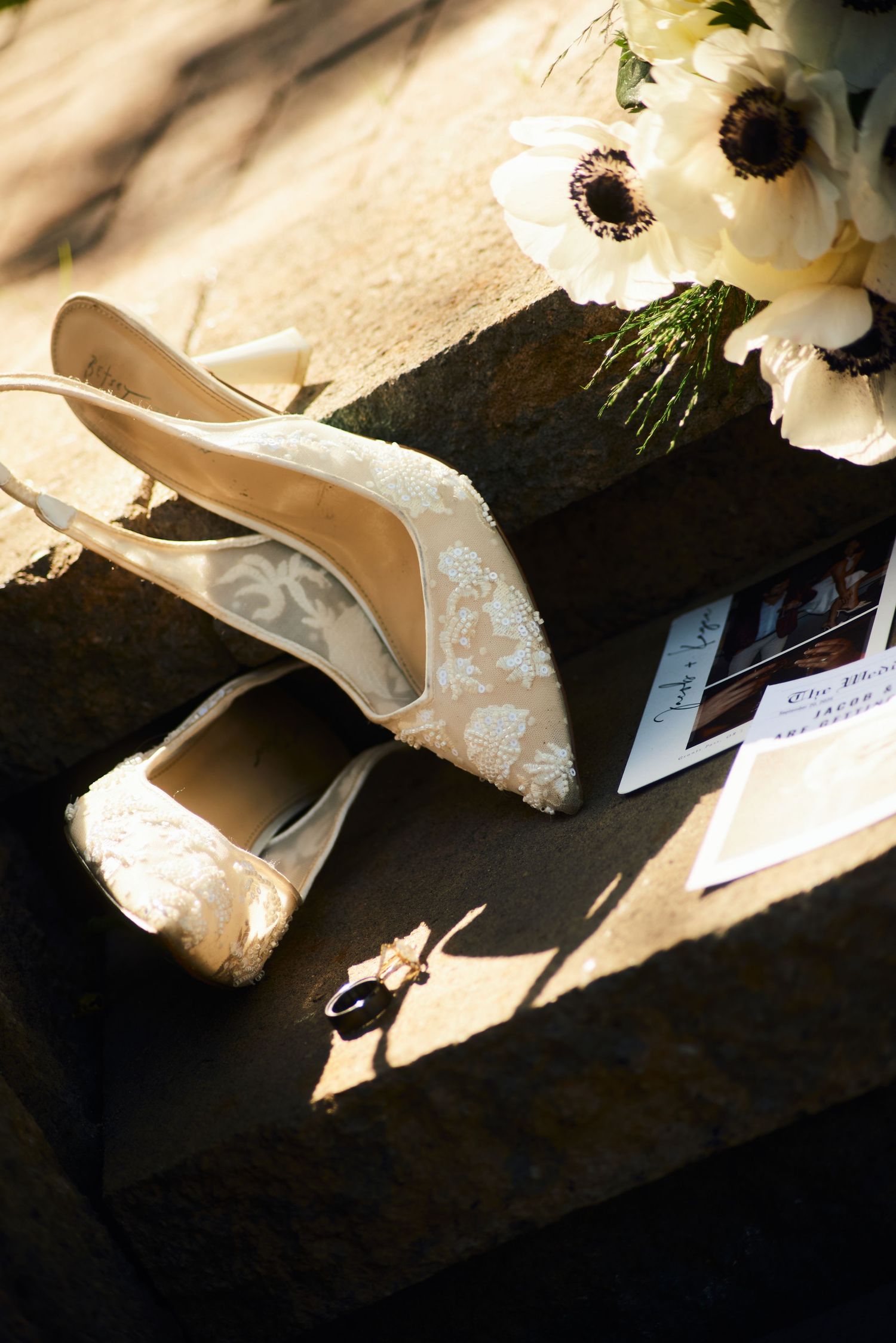 Elegant lace bridal heels displayed on stone steps beside white anemone bouquet, ring, and wedding cards in soft sunlight.