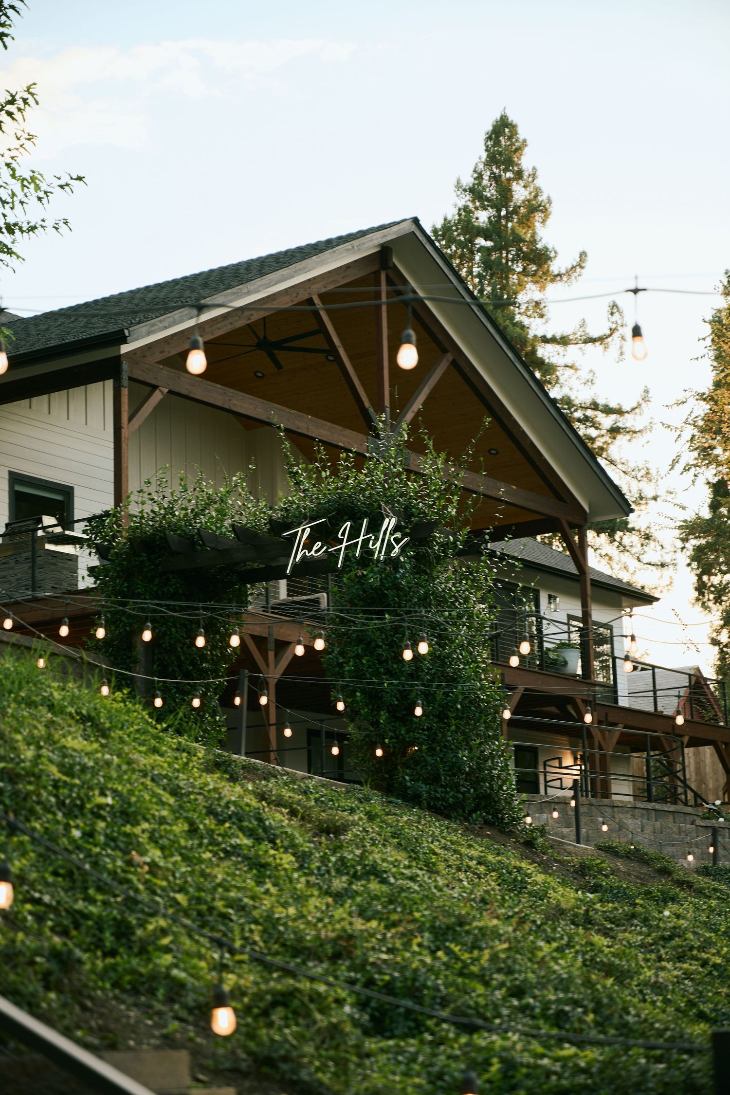 A rustic hillside restaurant with string lights and neon sign 'The Hub' glowing at dusk among green foliage.