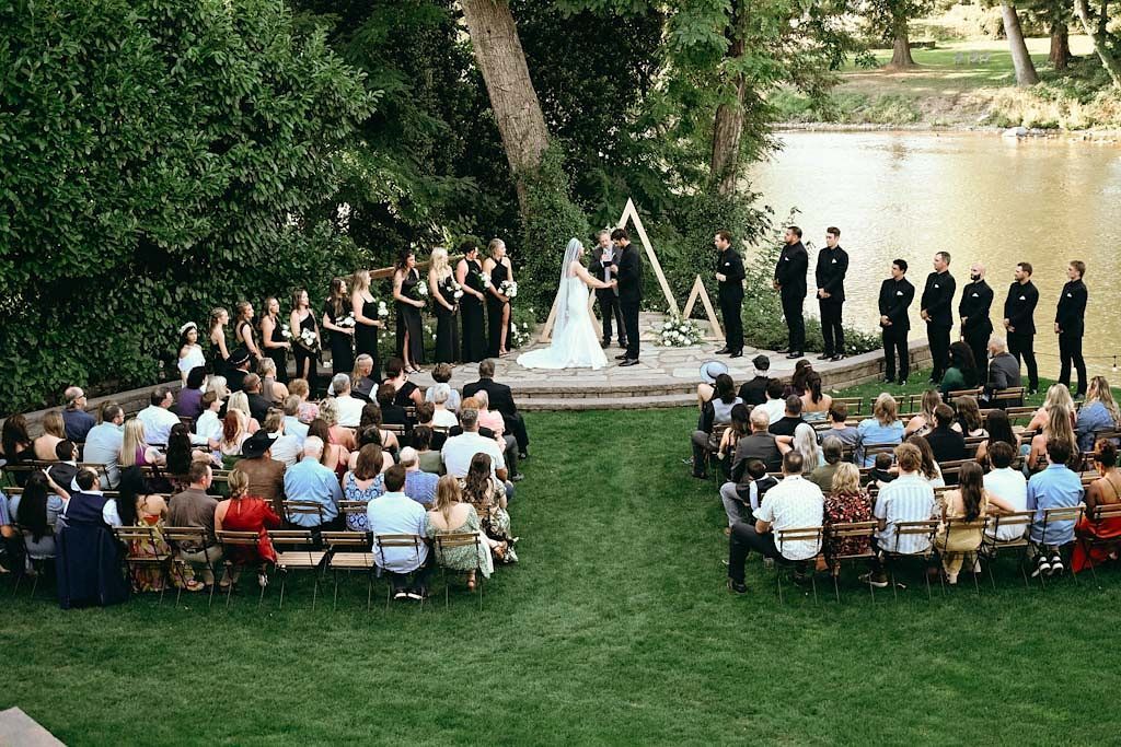 Outdoor wedding ceremony setup with rows of wooden chairs and guests under trees.