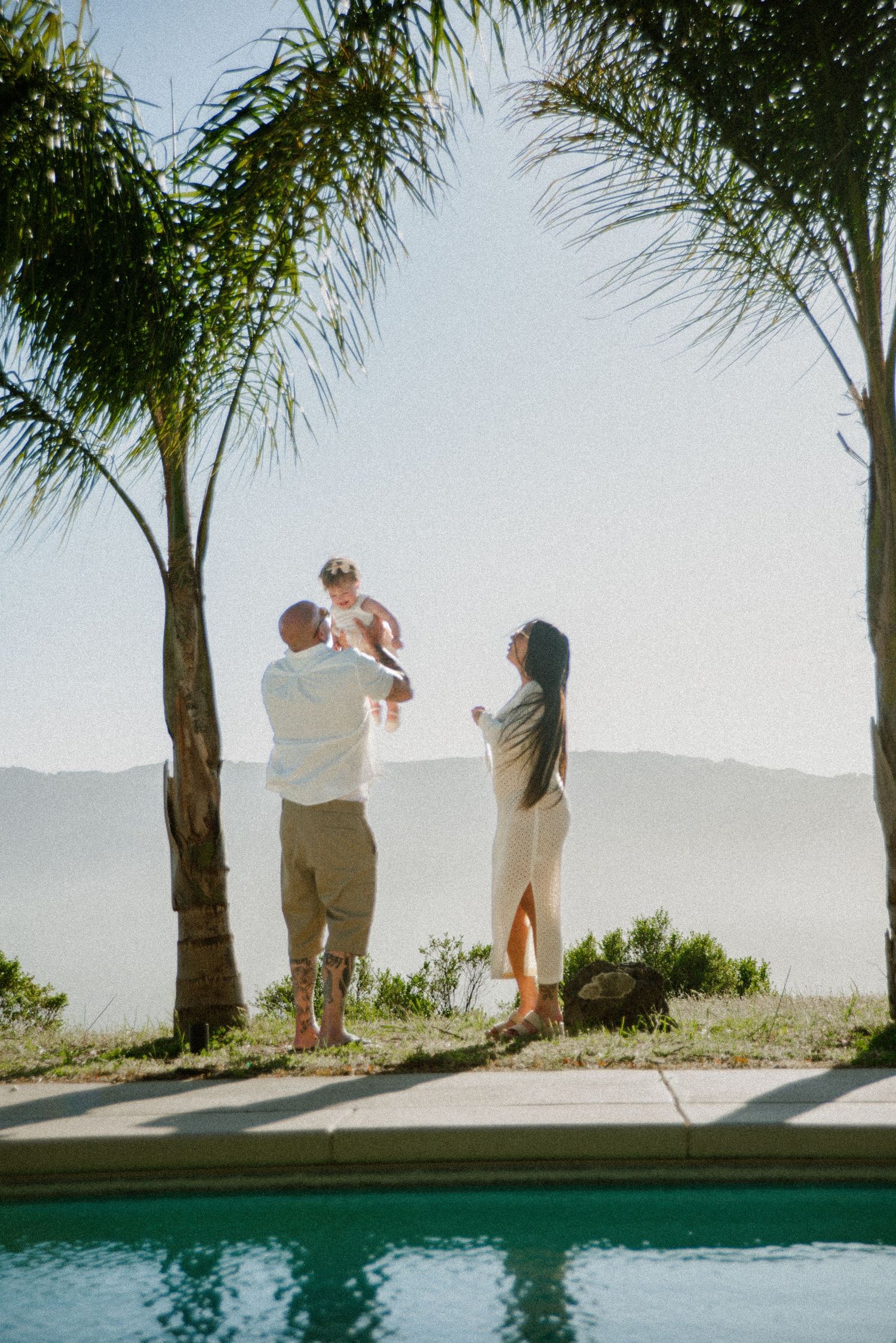Palm trees line a poolside as silhouettes move against a bright mountain backdrop.