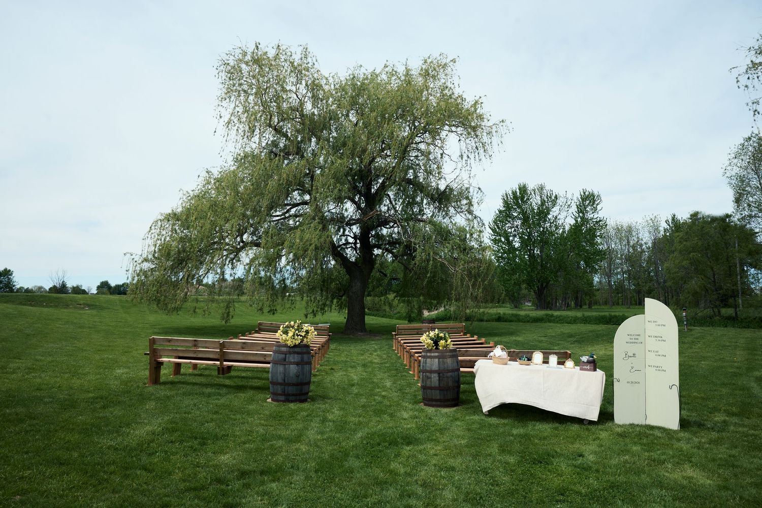 Rustic outdoor wedding ceremony setup with wooden barrels, benches, and a draped table beneath a weeping willow tree.