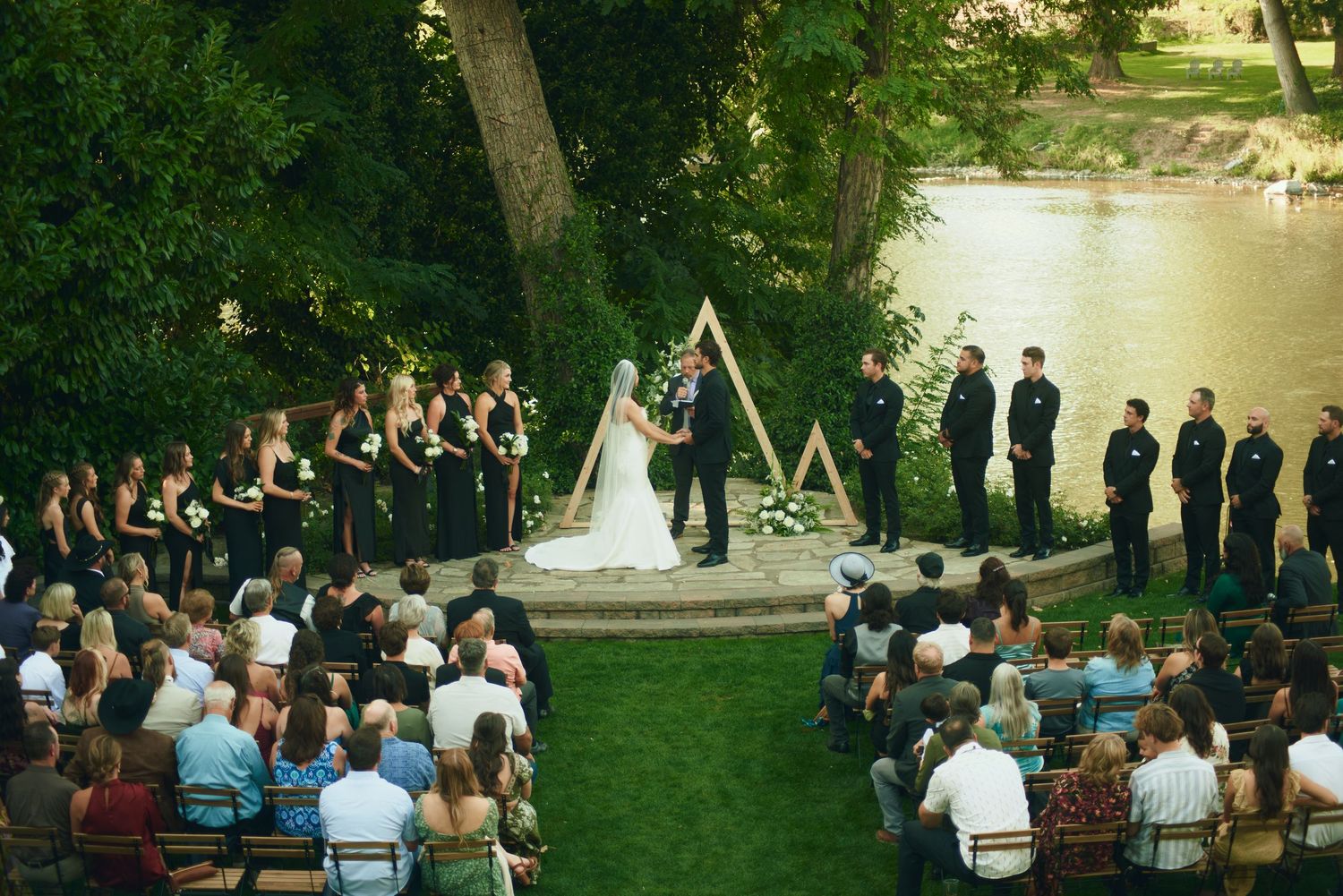 Outdoor wedding ceremony by riverside with bridal party in black, guests seated, and triangular arch altar on green lawn.