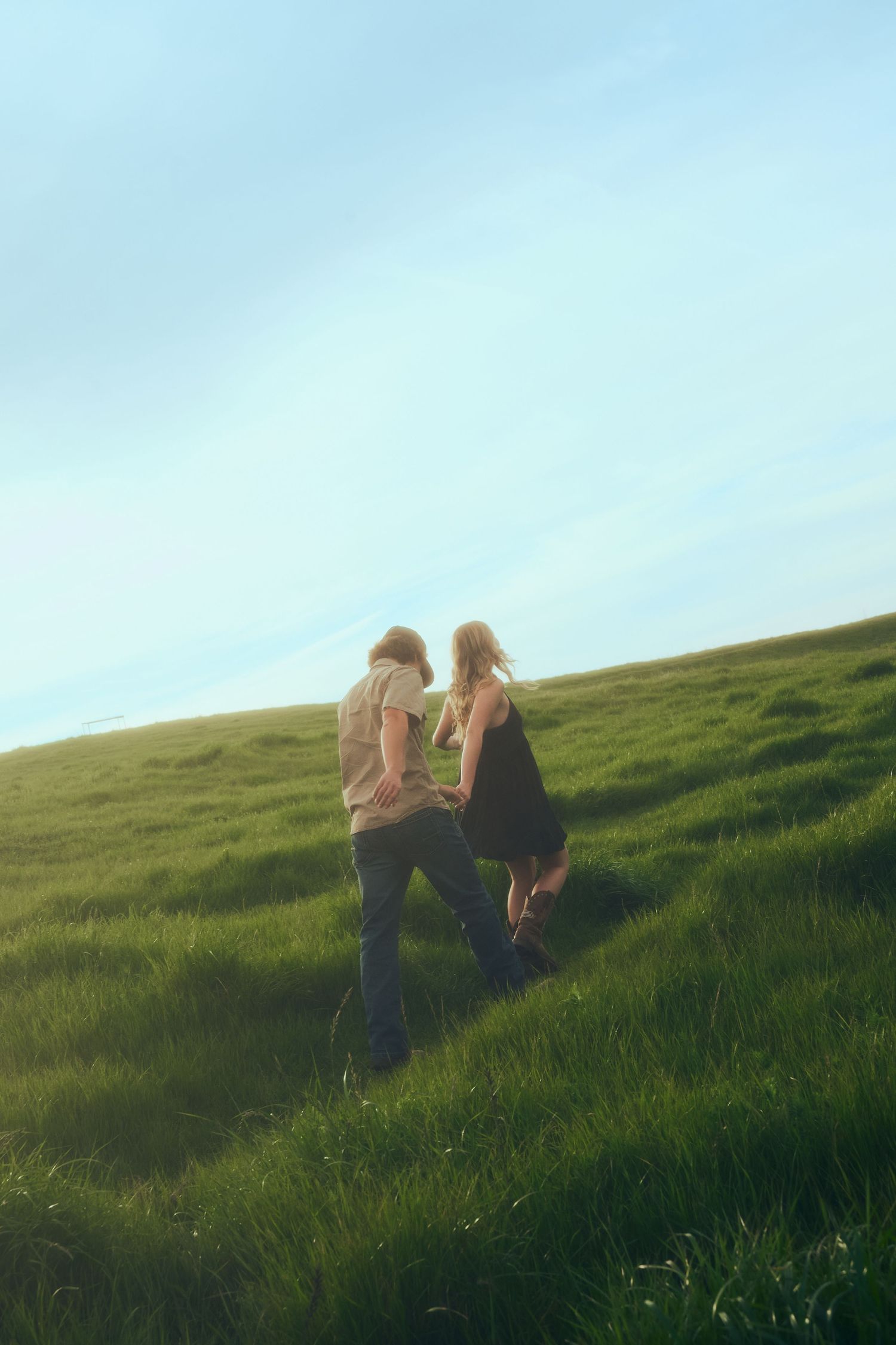 A couple walks hand-in-hand up a lush green hillside meadow under a bright blue sky.