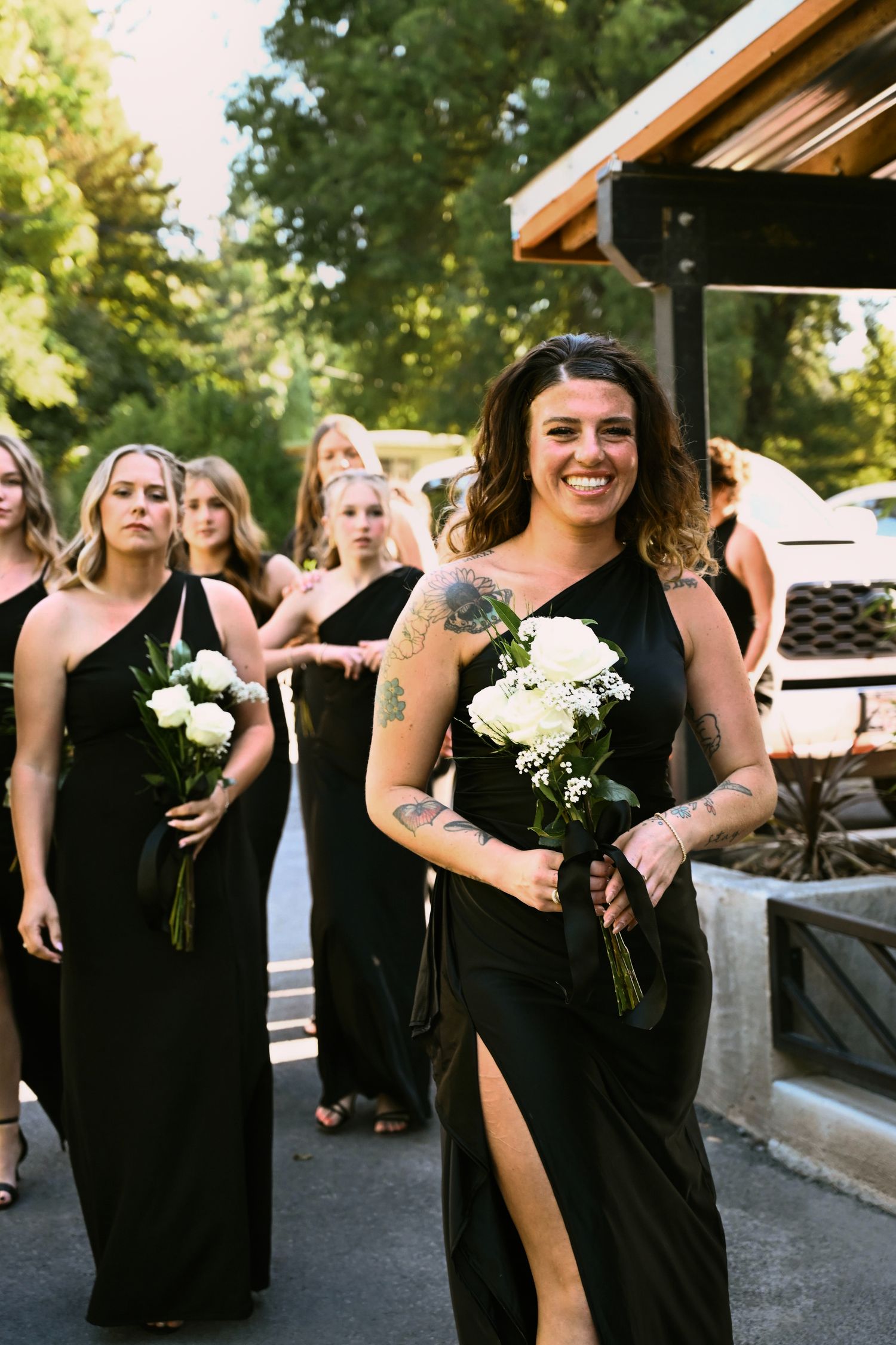 Smiling bridesmaids in black gowns carry white bouquets while walking outdoors at a wedding ceremony.
