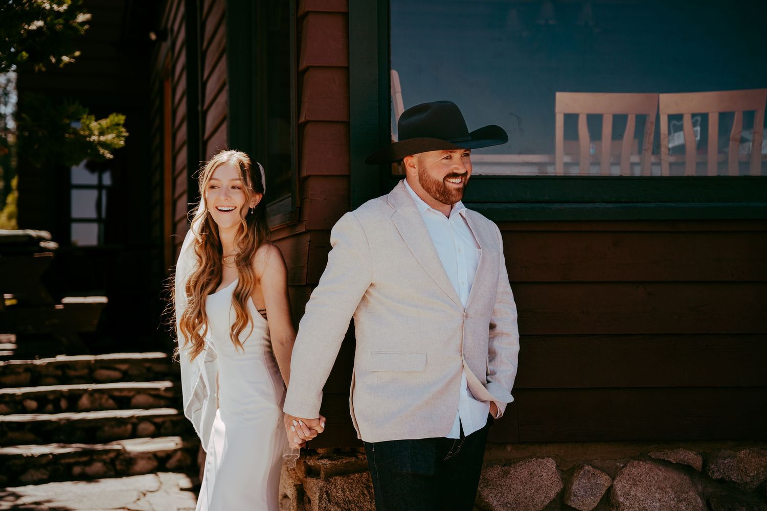 Bride in white gown and groom in beige suit with cowboy hat hold hands outside rustic wooden building.