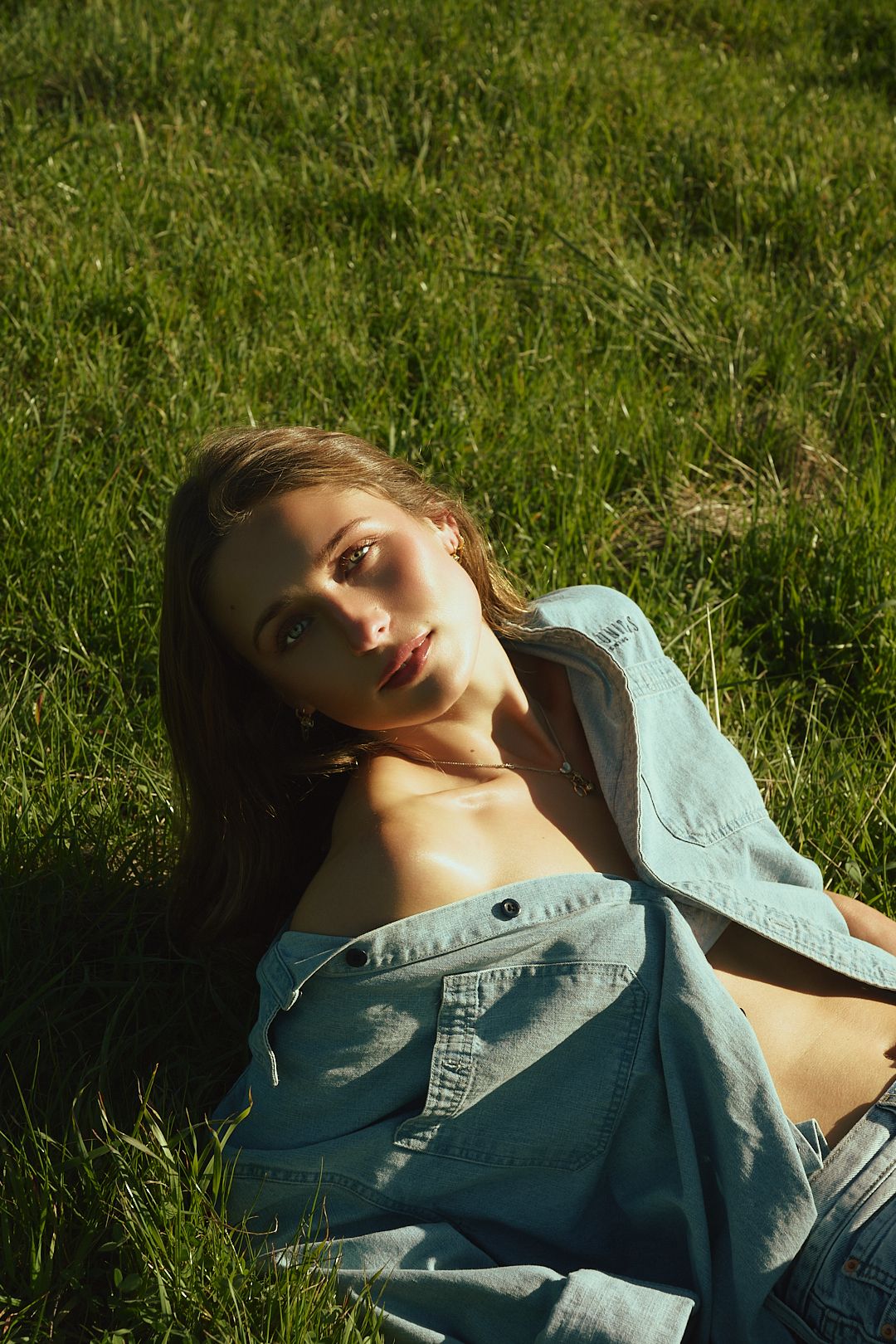 Young woman in denim shirt lying on green grass in sunlight, looking up with a relaxed expression.