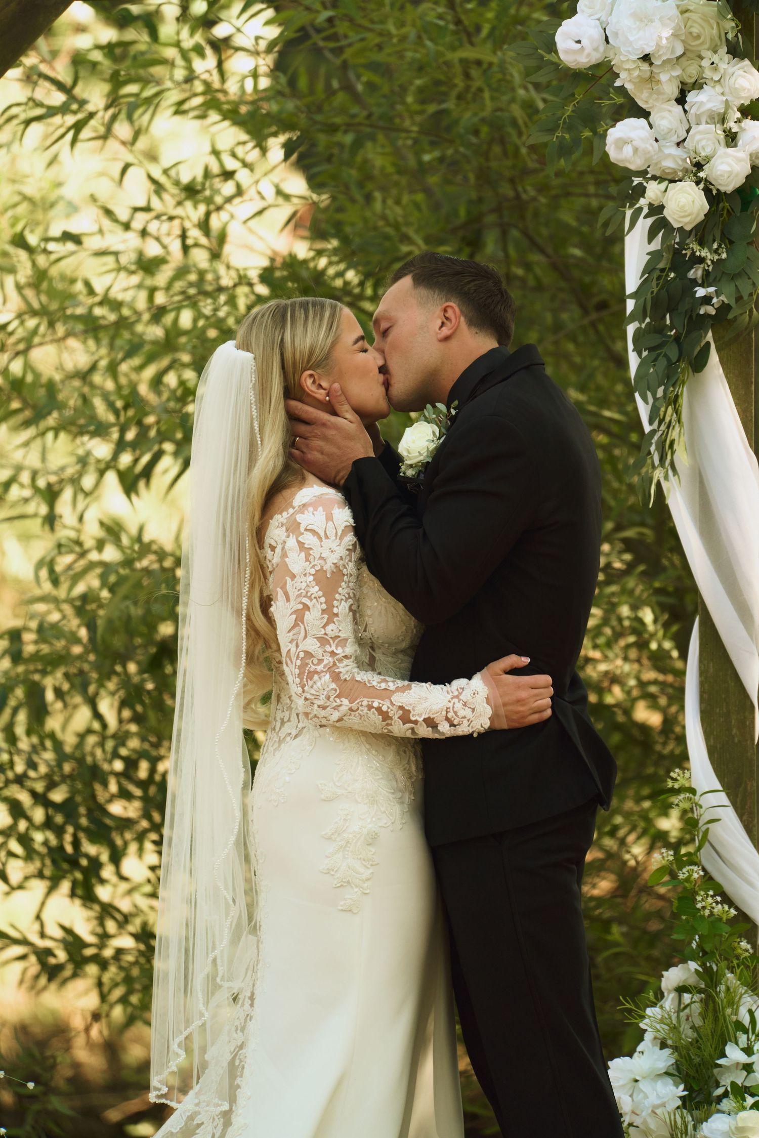 Bride and groom share first kiss at outdoor wedding ceremony beneath floral arch surrounded by greenery.