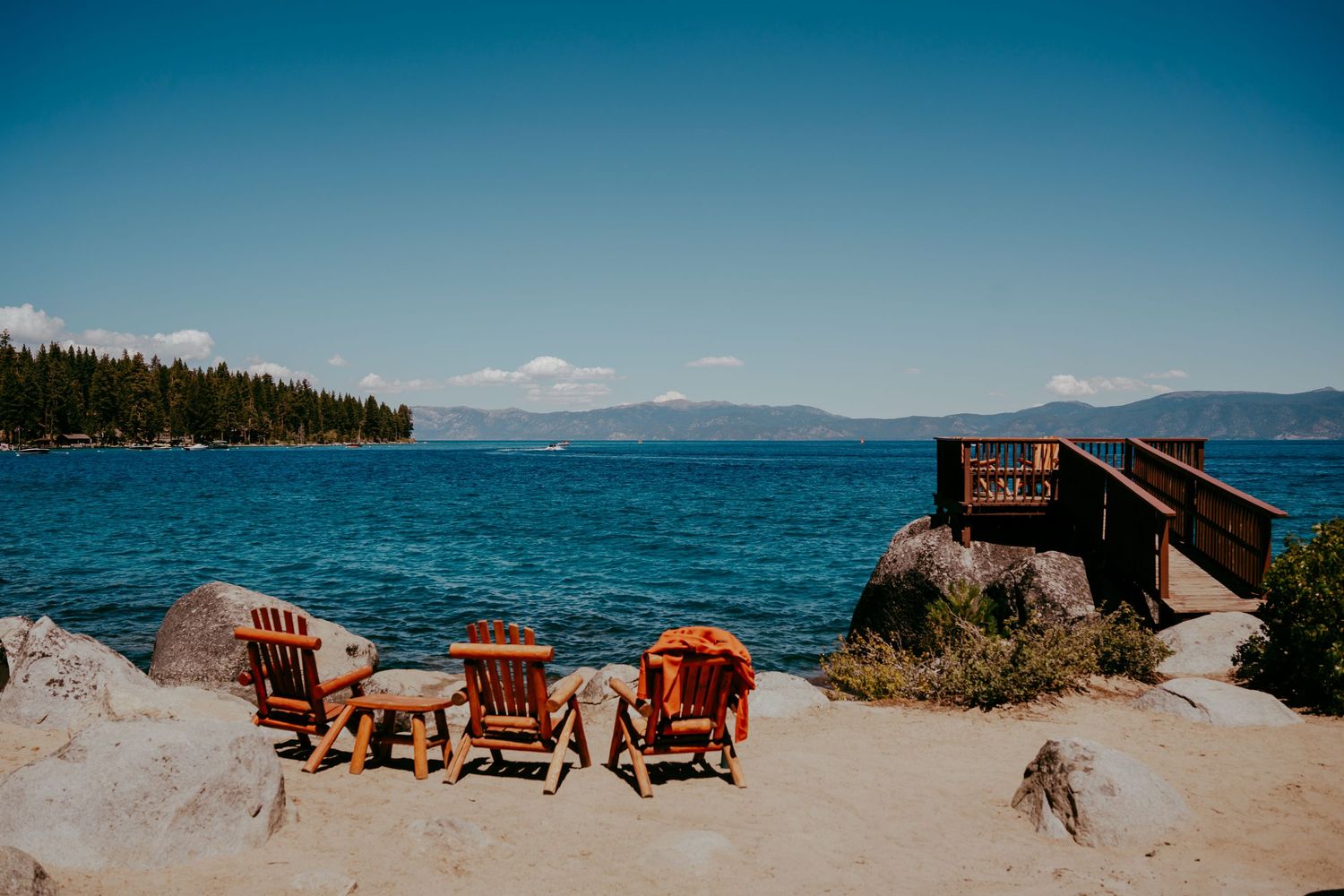 Wooden Adirondack chairs on sandy Lake Tahoe shore with turquoise water, pine trees, and mountains under blue sky.
