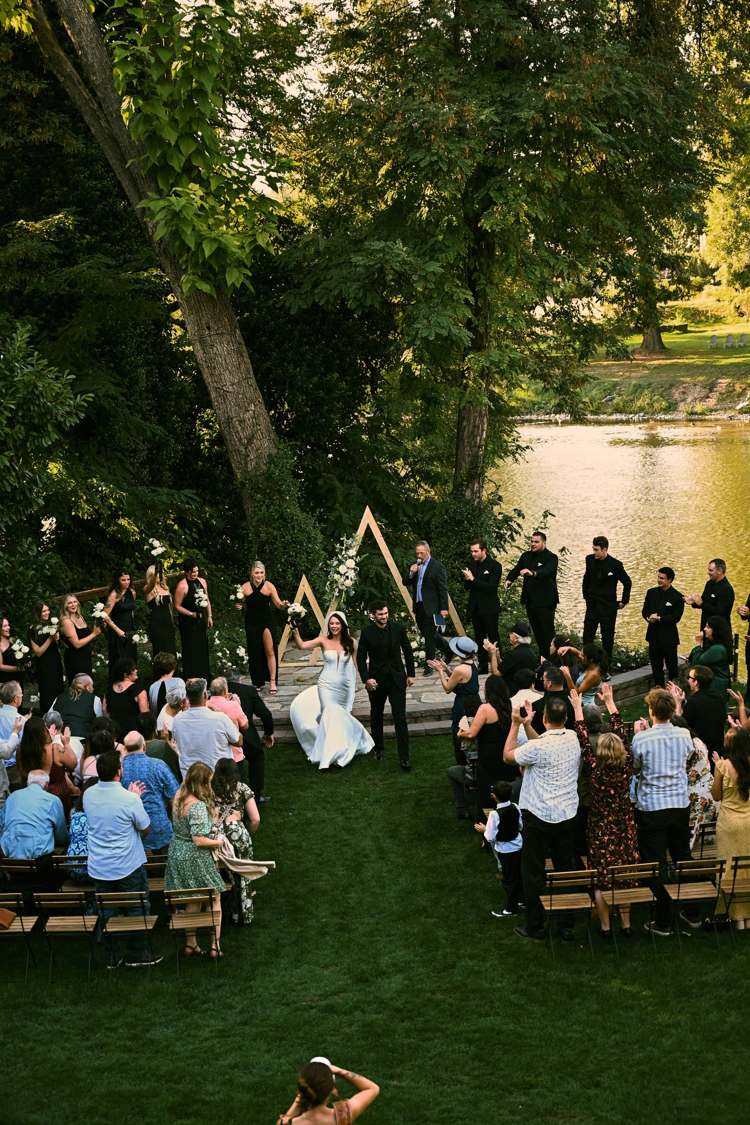 Aerial view of an outdoor wedding ceremony by a lake, with guests seated on wooden benches as the bride and groom walk the aisle.