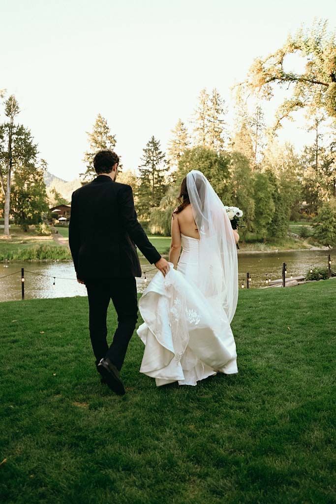 Newlyweds walk hand in hand across a grassy lawn with a lake and trees in the background.