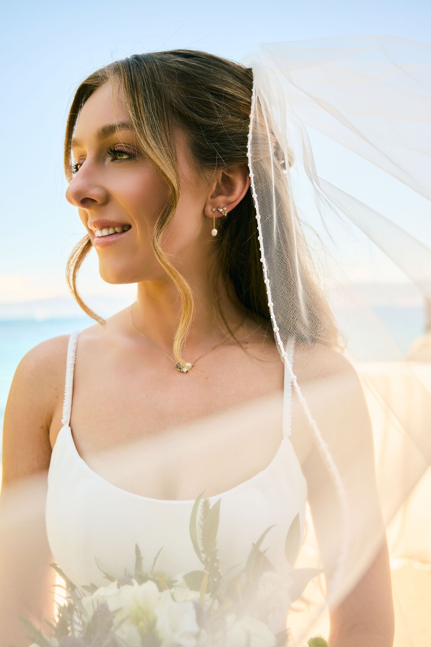 Smiling bride in white dress and veil holding bouquet on beach with blue ocean backdrop.