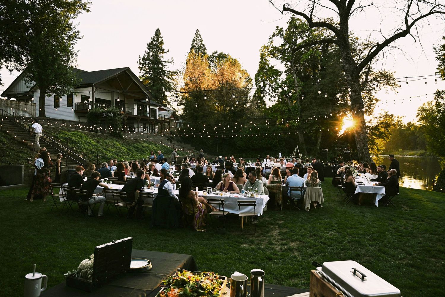 Outdoor wedding reception at sunset with guests seated at white-draped tables on a lush lawn with string lights.