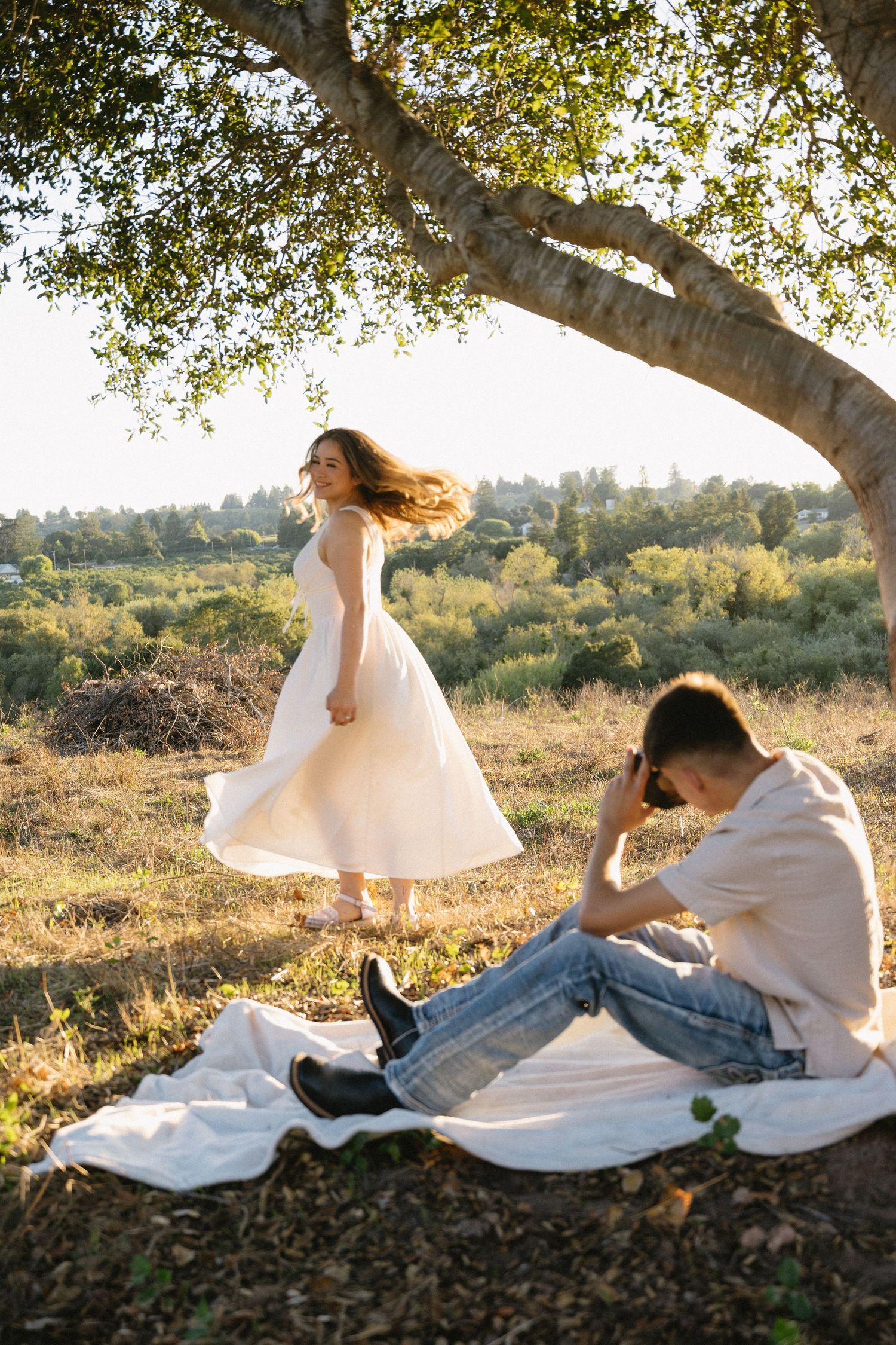 Romantic couple's photo session under tree with flowing white dress and natural lighting.