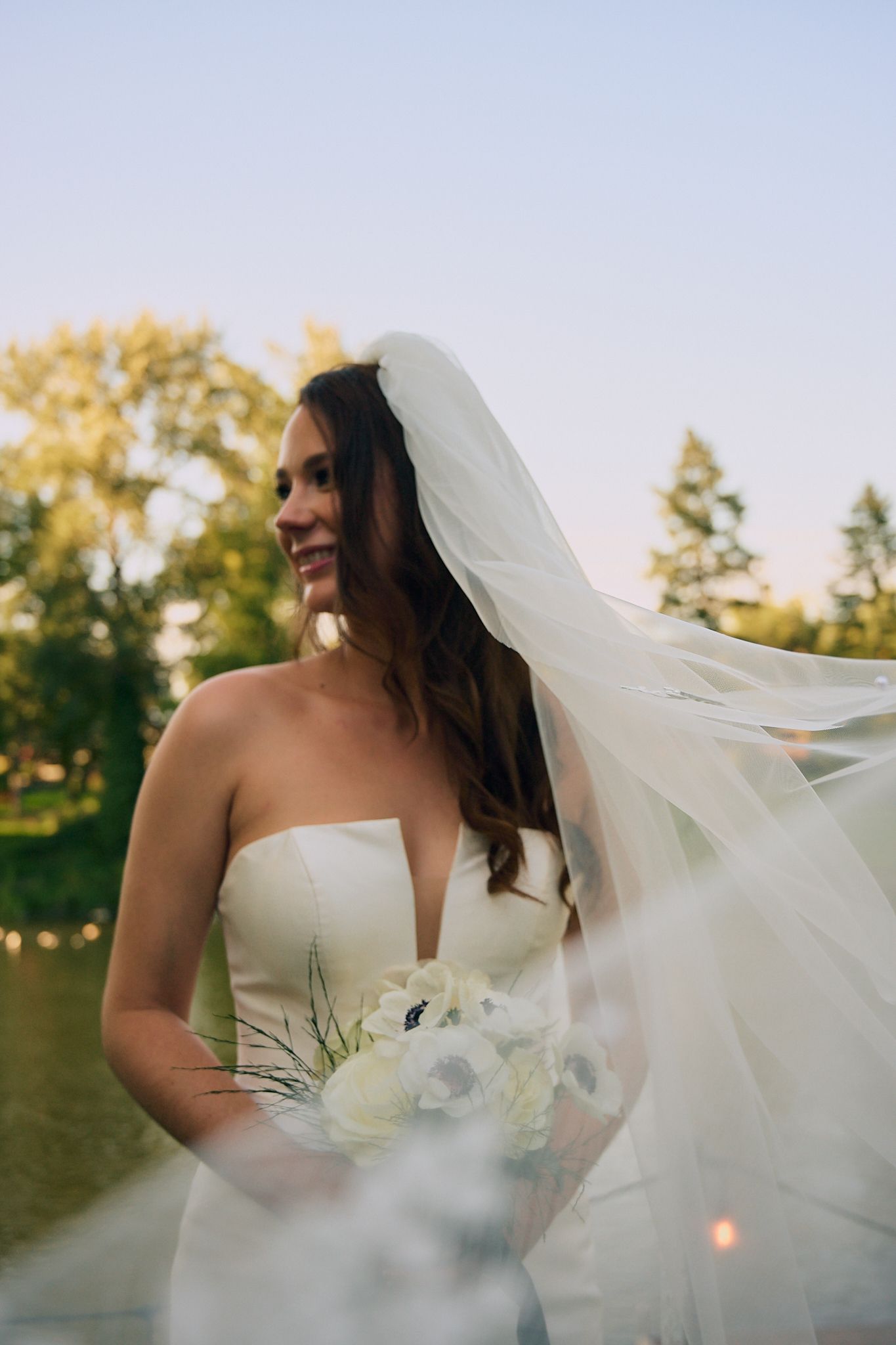 Smiling bride in strapless gown holding white bouquet, veil flowing in breeze near lake at golden hour.