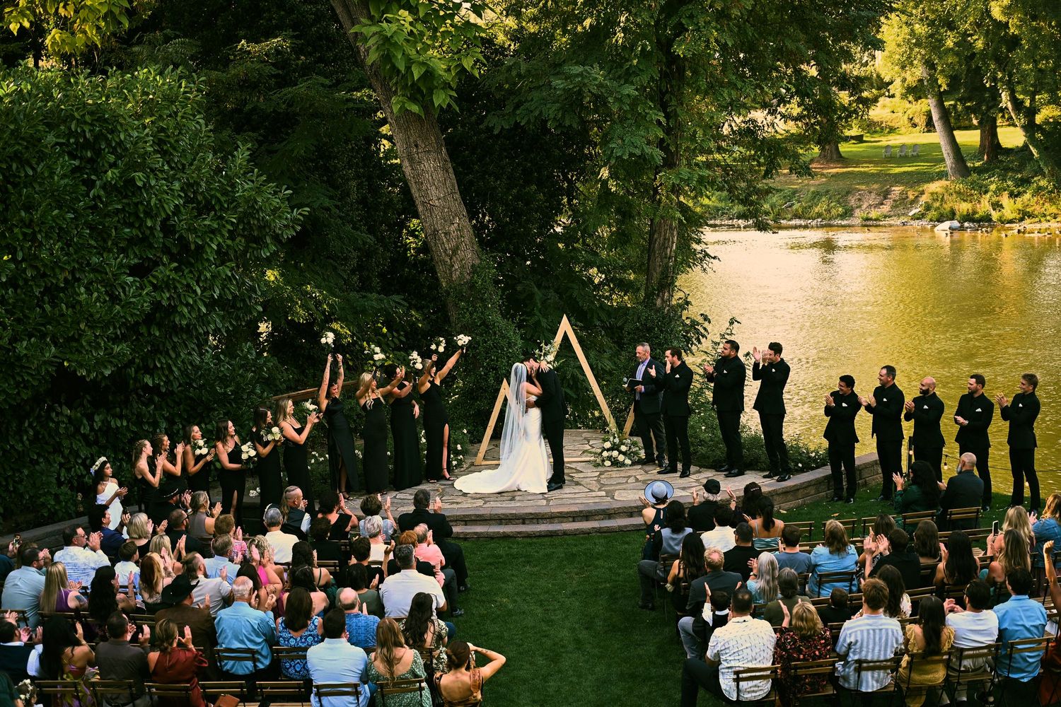 Outdoor wedding ceremony by a river with bride, groom, wedding party, and seated guests surrounded by lush green trees.