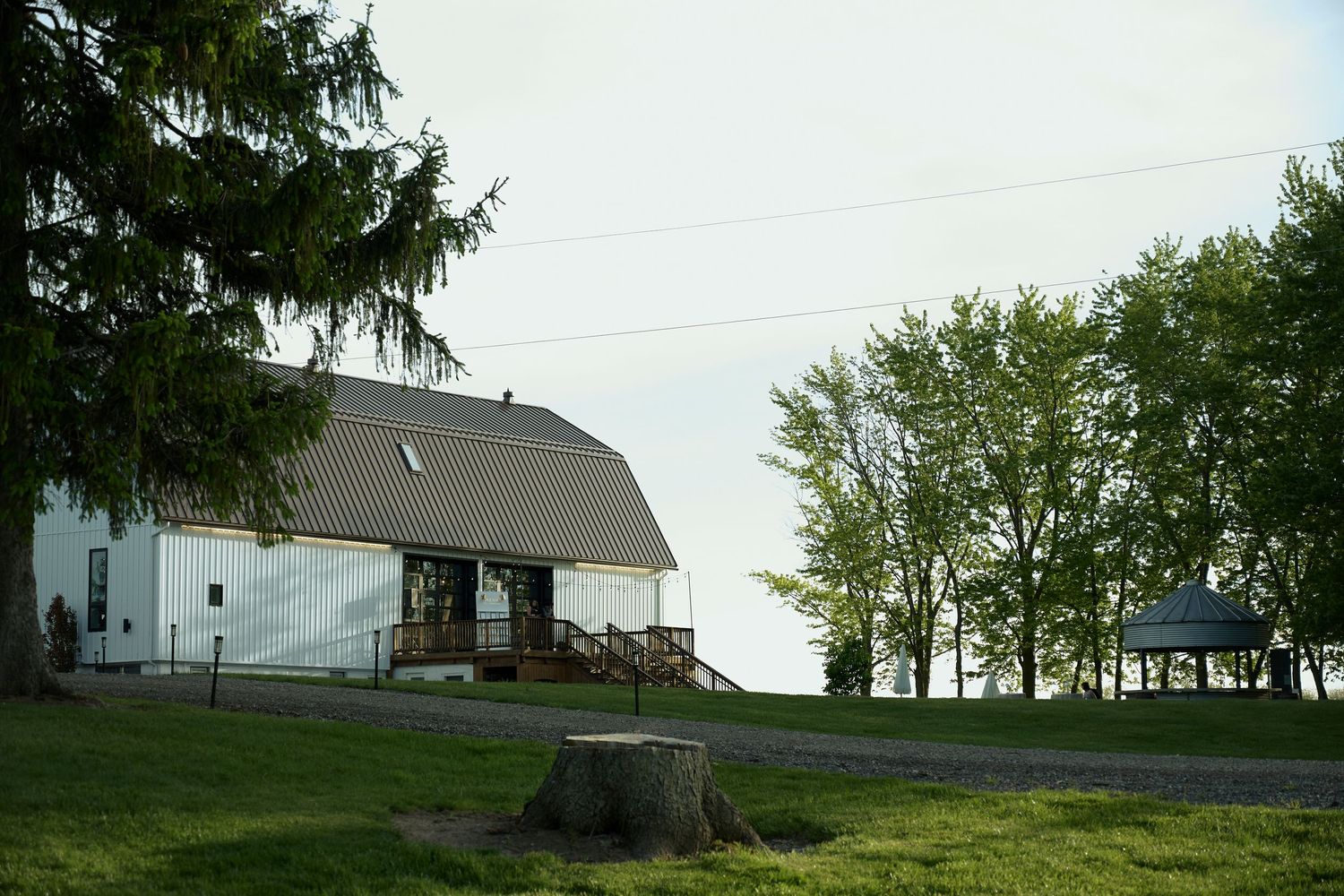 White barn with gambrel roof on a green hill surrounded by tall trees, with a tree stump in the foreground.