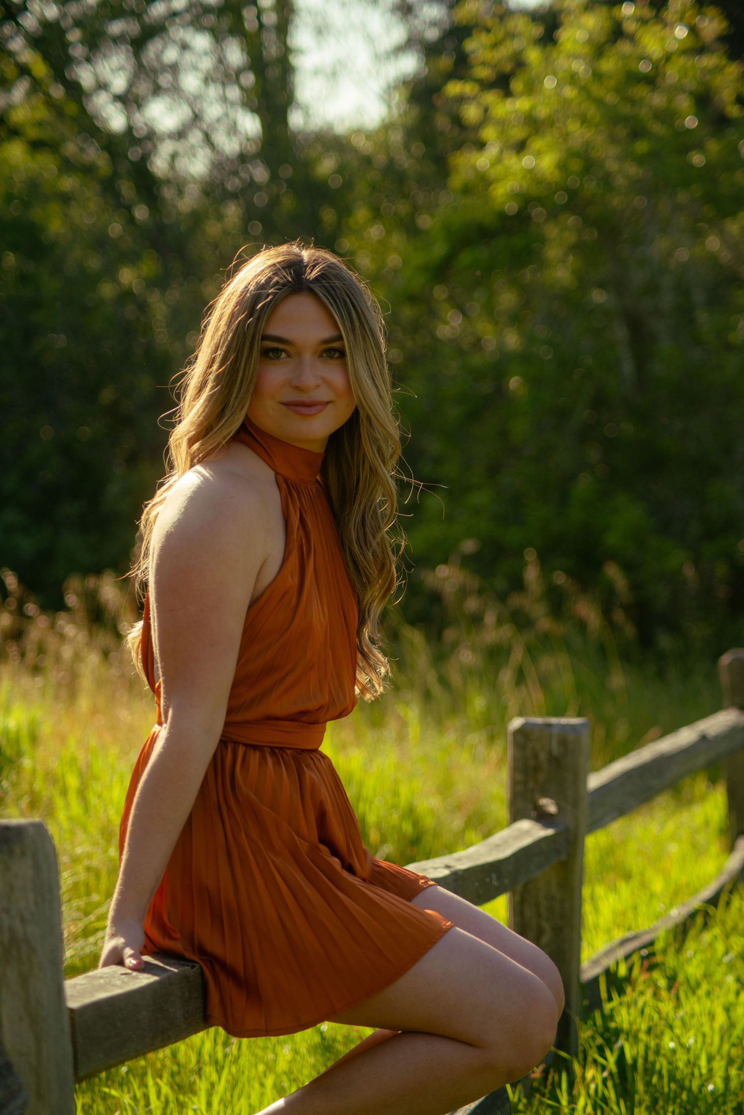 A person in a rust dress poses gracefully by a wooden fence in a grassy field at sunset.