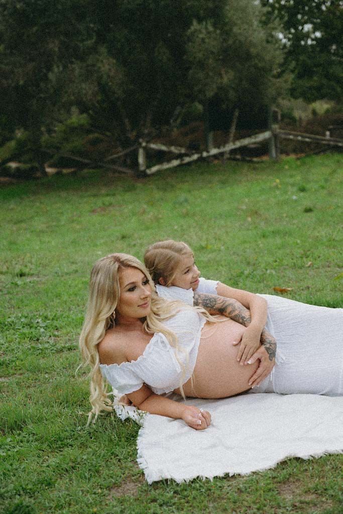A pregnant woman in white dress lounges with a child on a blanket outdoors.