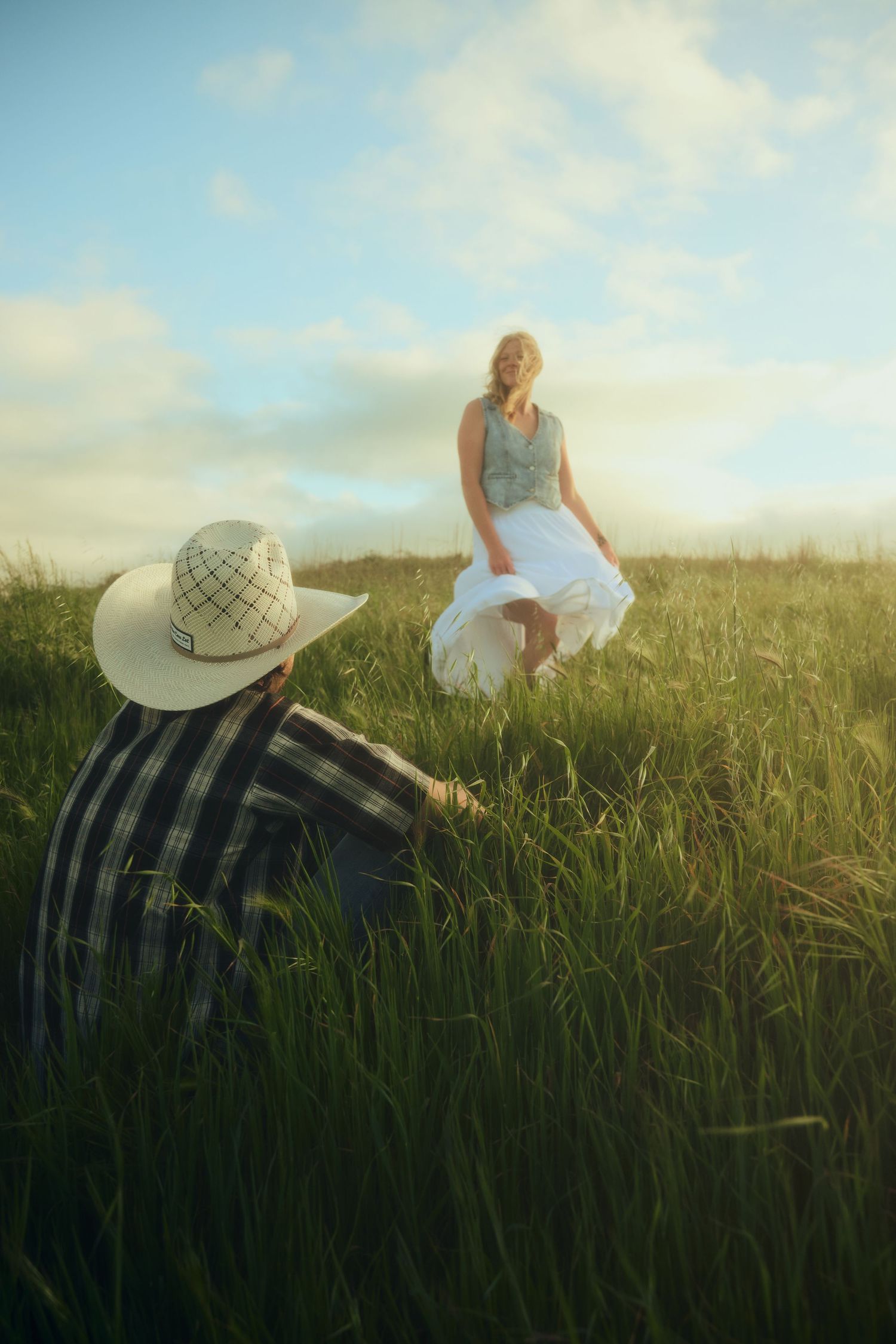 A woman in a white dress stands in a green field while a man in a plaid shirt and cowboy hat sits nearby.