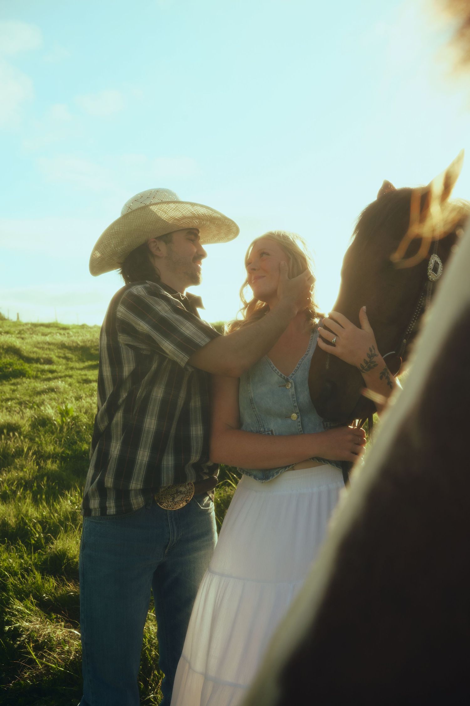 Couple with cowboy hat embracing near a brown horse in a sunlit green field.