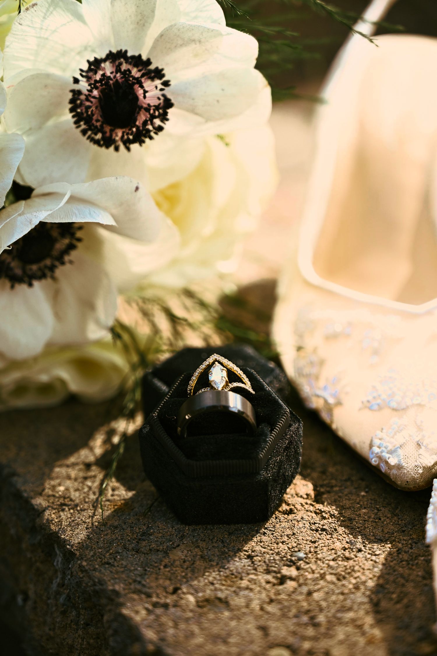 Black velvet ring box with diamond engagement ring and wedding band beside white anemone flowers and embellished bridal heels.