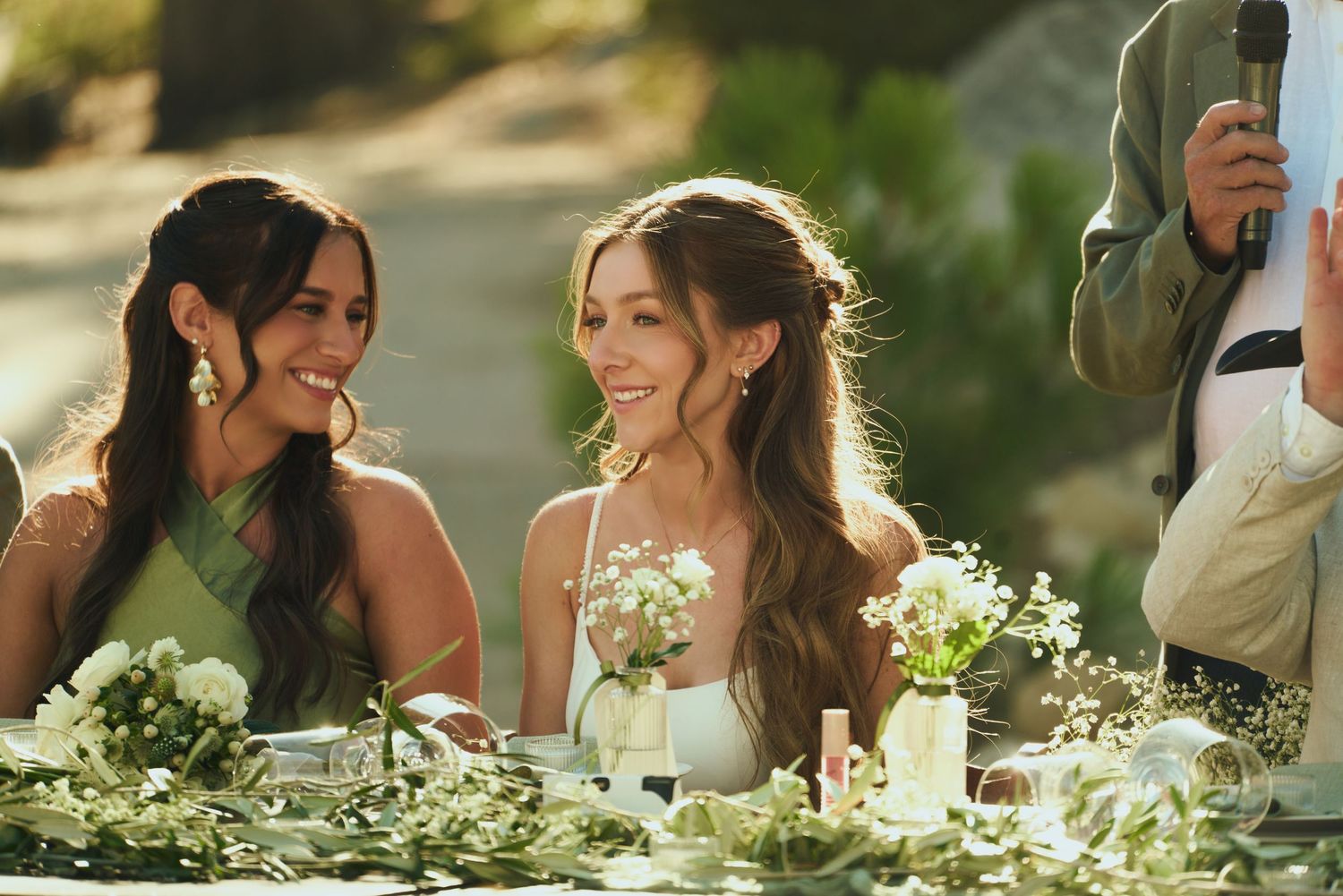 Two bridesmaids in elegant attire smile and laugh at a floral-decorated outdoor wedding reception table.