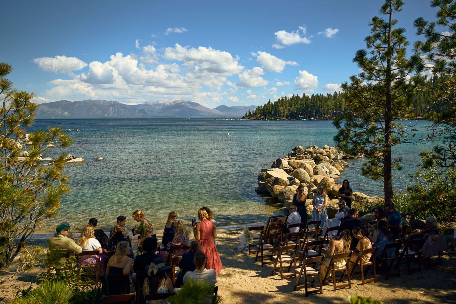 Outdoor wedding ceremony by Lake Tahoe shore with guests seated among pine trees, turquoise water and mountains in background.