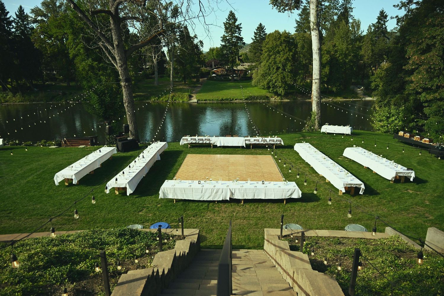 Outdoor wedding reception setup with long white tables on green lawn beside a serene pond surrounded by tall trees.
