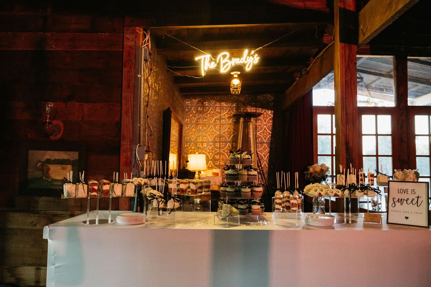 Rustic wedding dessert table with neon sign, floral arrangements, and sweet treats in a barn venue.