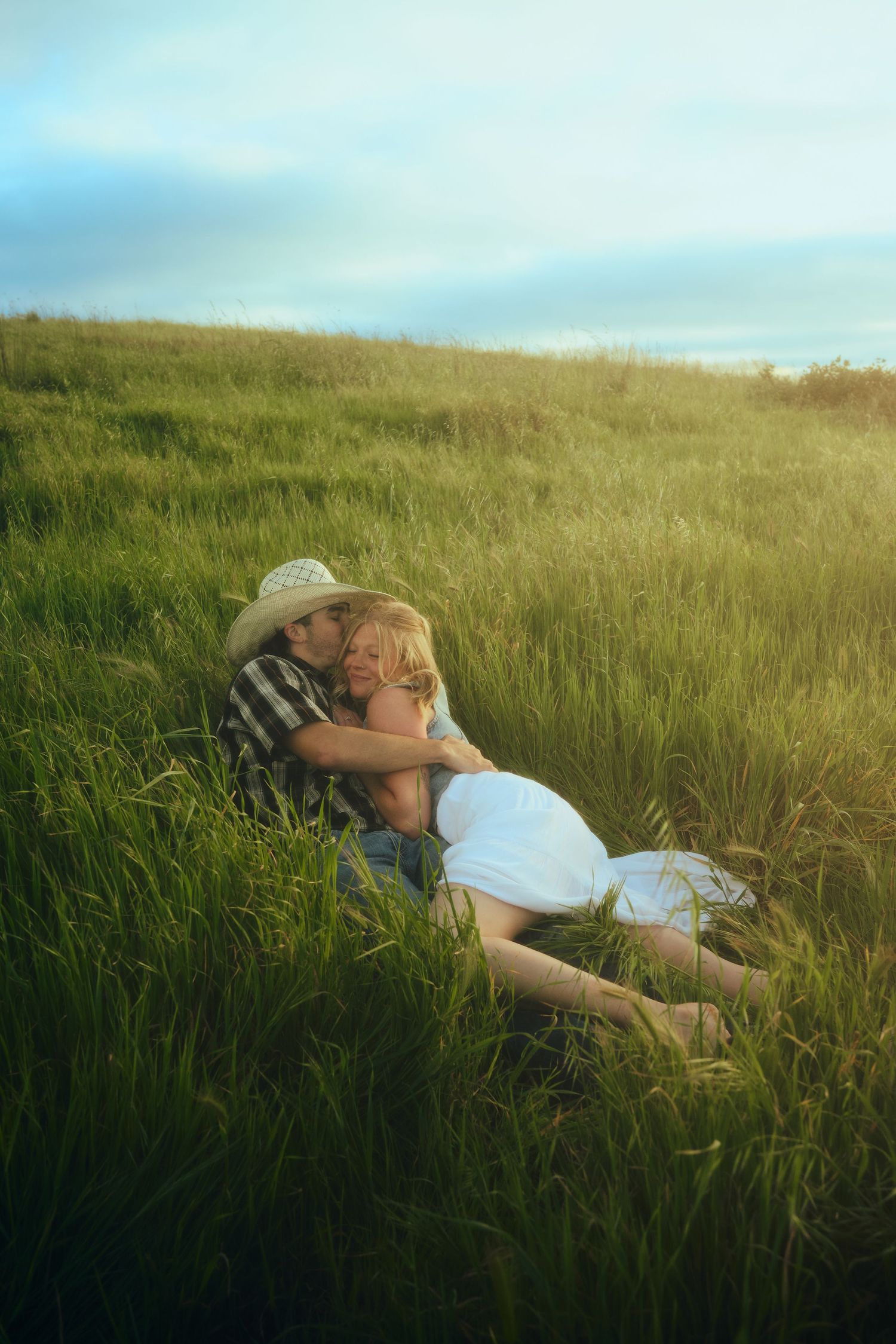 Romantic couple embracing in a lush green field at golden hour, man in cowboy hat kissing woman in white dress.