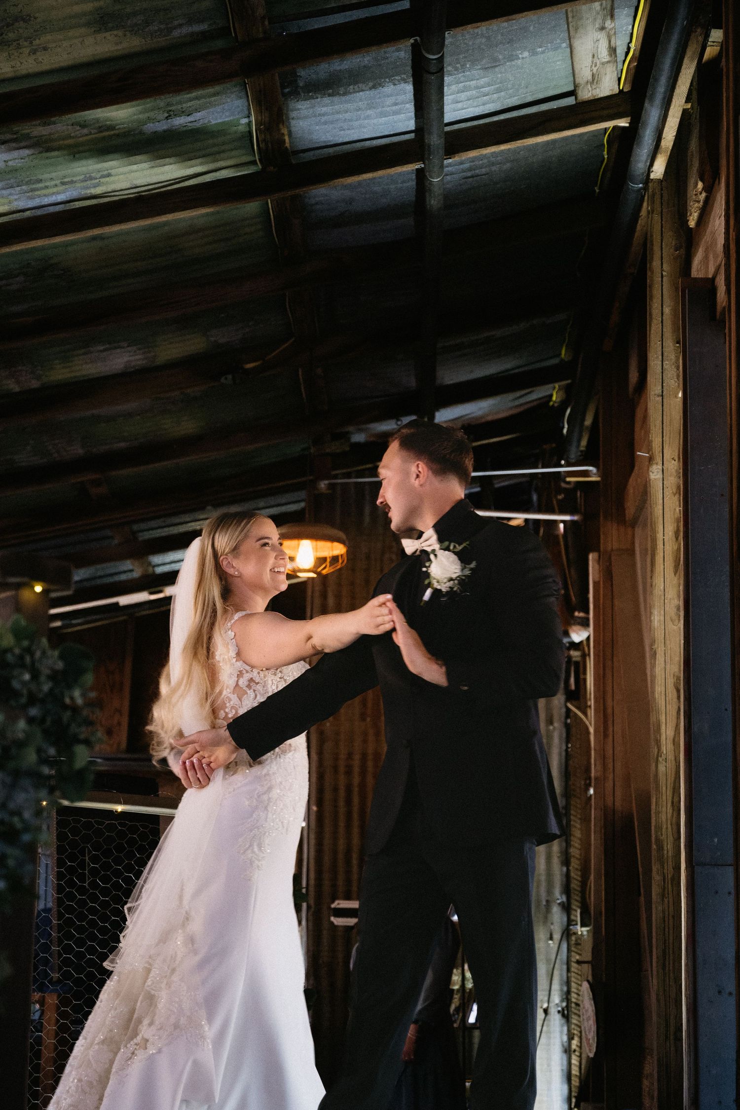 Bride and groom share first dance in rustic barn venue, bride in lace gown, groom in black suit with boutonniere.