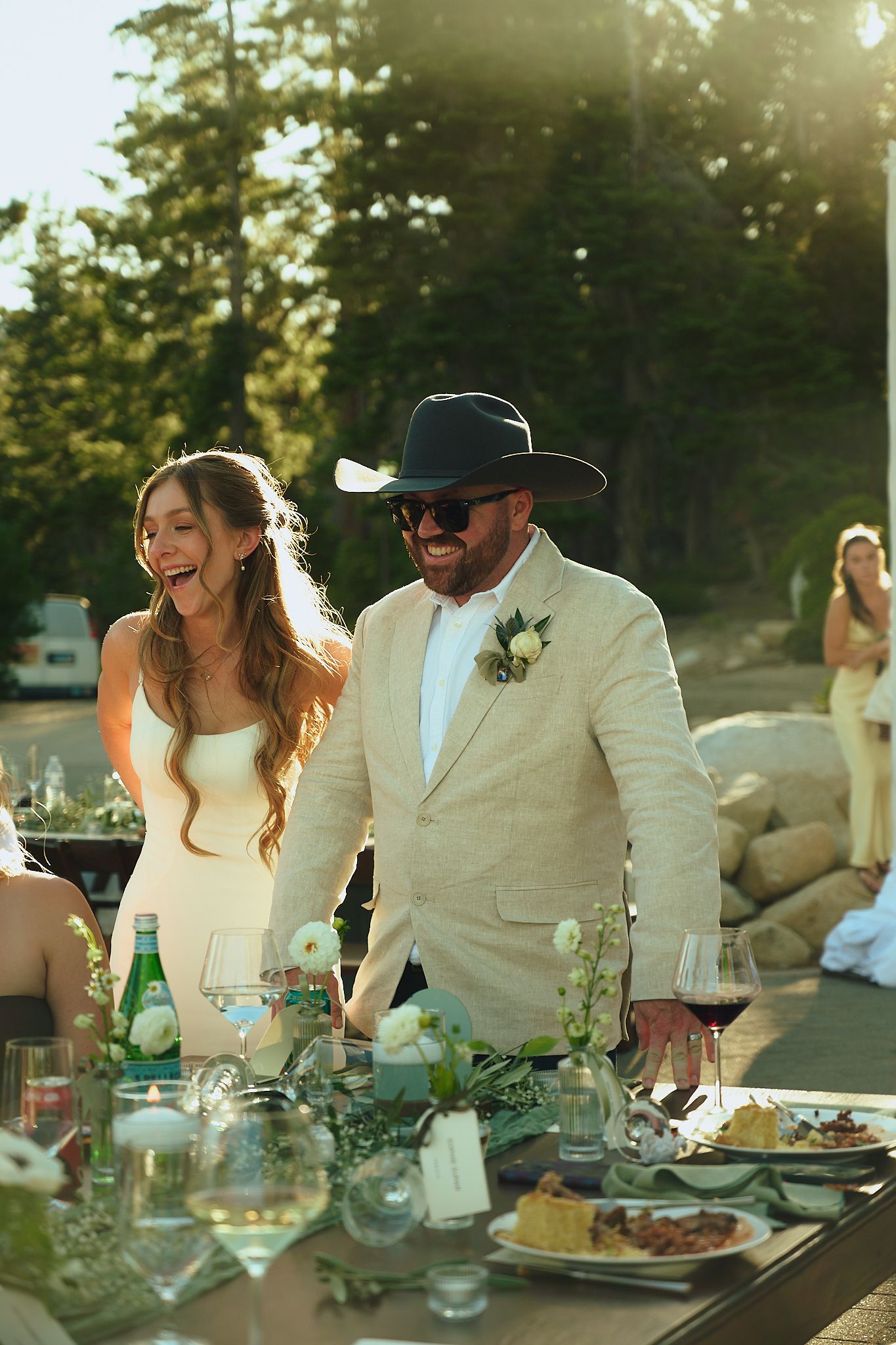 Bride and groom laughing together at outdoor wedding reception table, groom wearing tan suit and black cowboy hat.