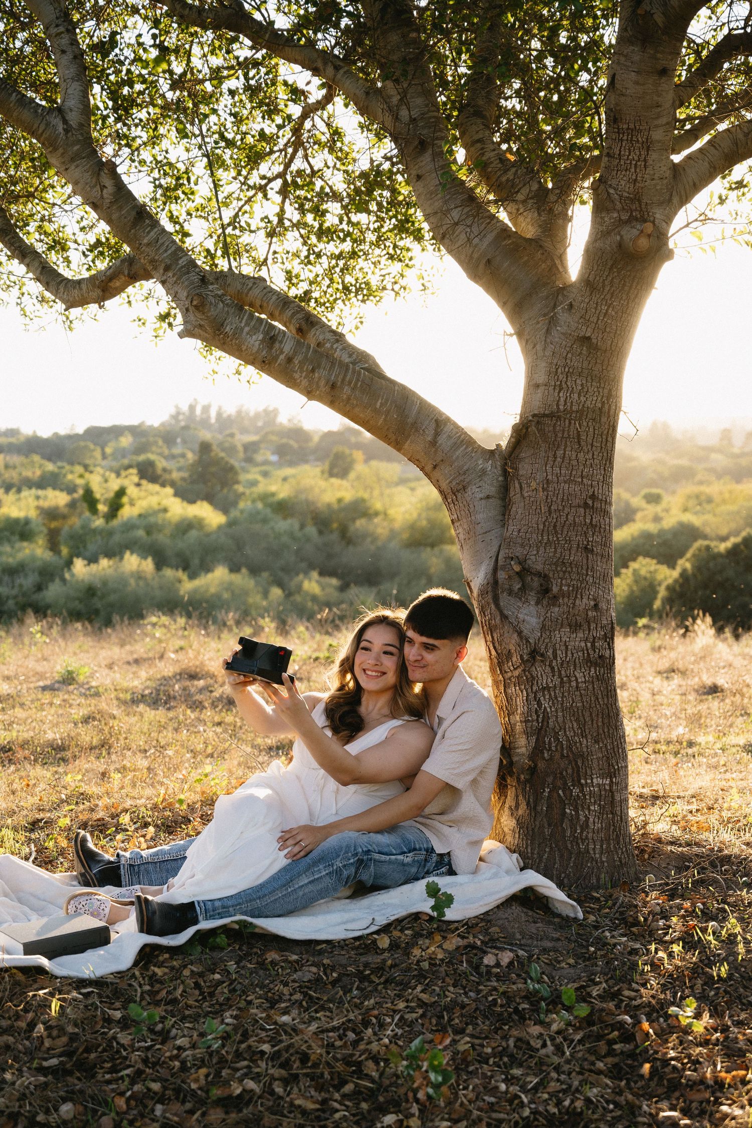 Couple relaxing under large tree during sunset photoshoot.