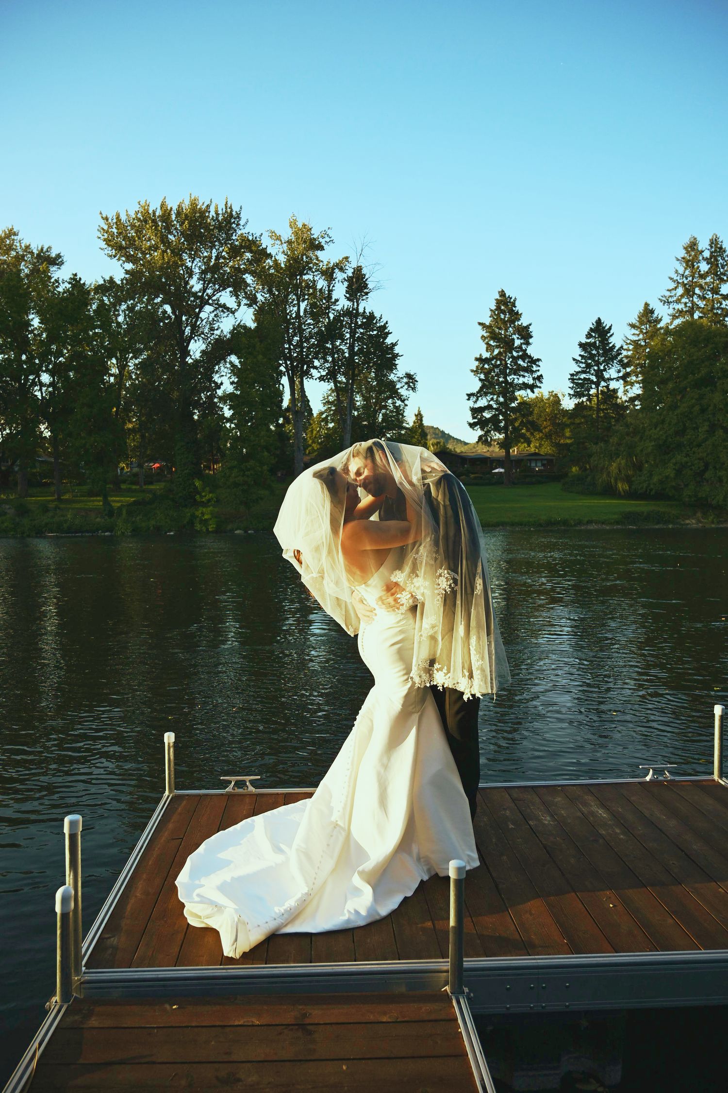 Bride and groom share romantic moment under veil on wooden dock by serene lake at golden hour.