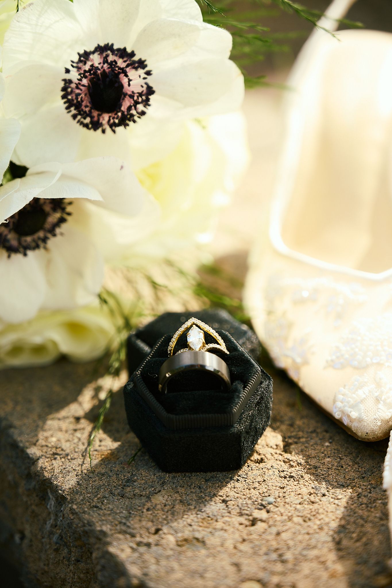 Black velvet ring box with gold wedding bands beside white anemone flowers and lace bridal heel on stone.