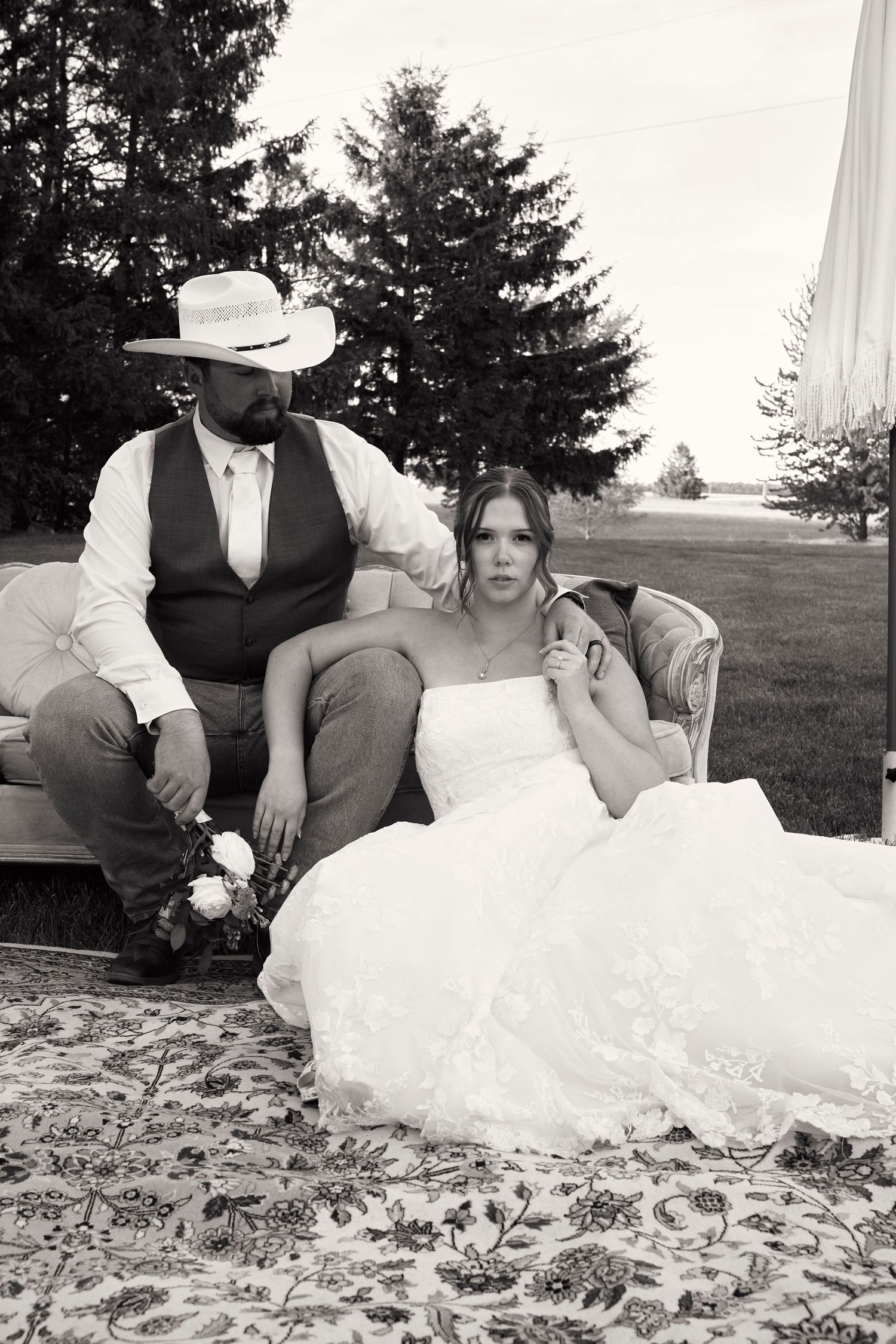 Black and white photo of a bride in a white gown and groom in a vest and cowboy hat seated outdoors on a vintage sofa.