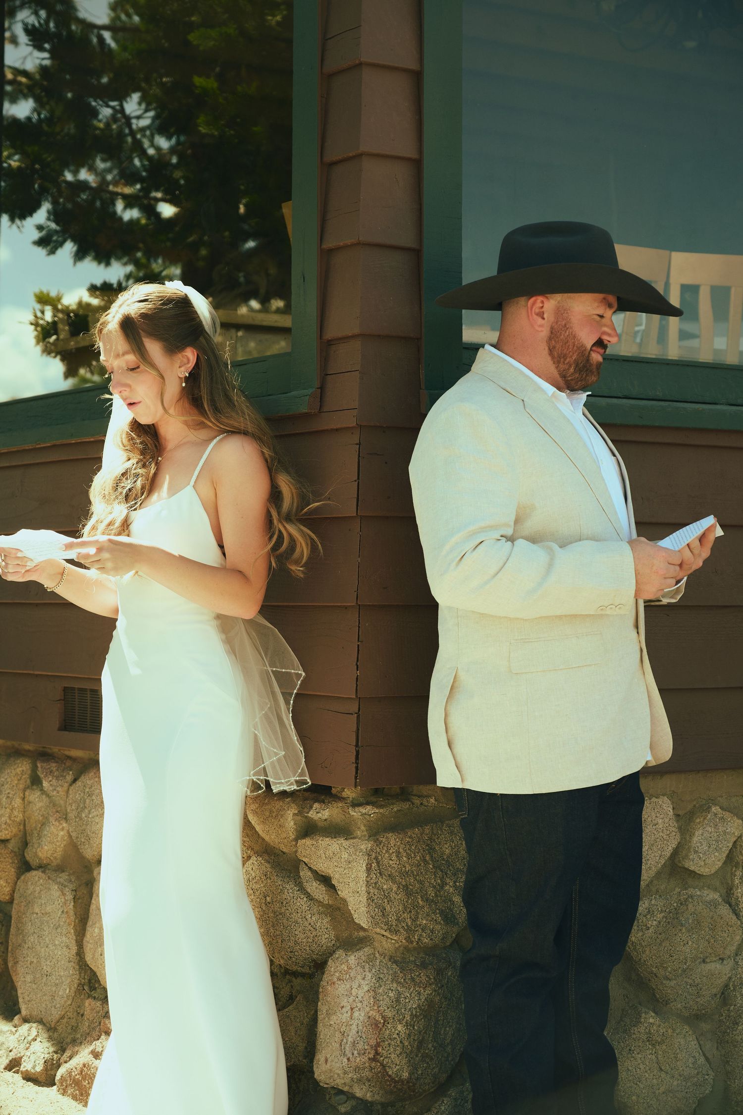 Bride in white gown and groom in cream suit with black cowboy hat reading vows back-to-back outdoors.