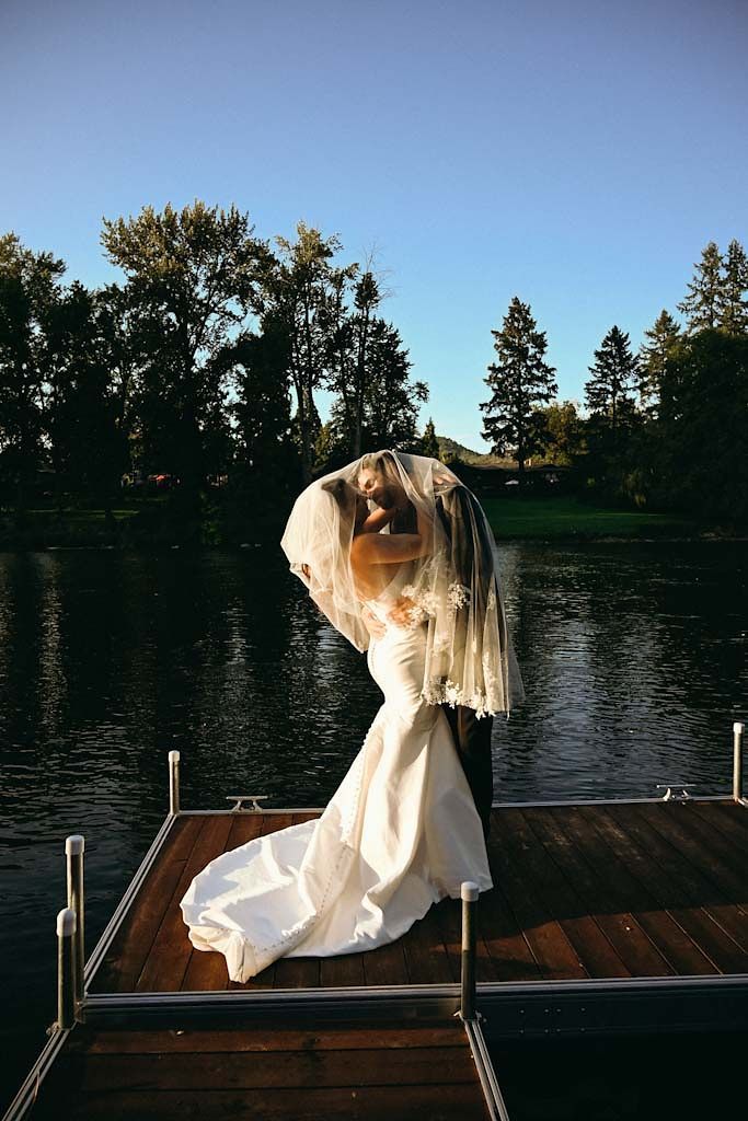 Bride sitting on wooden dock by lake during sunset in flowing white dress.