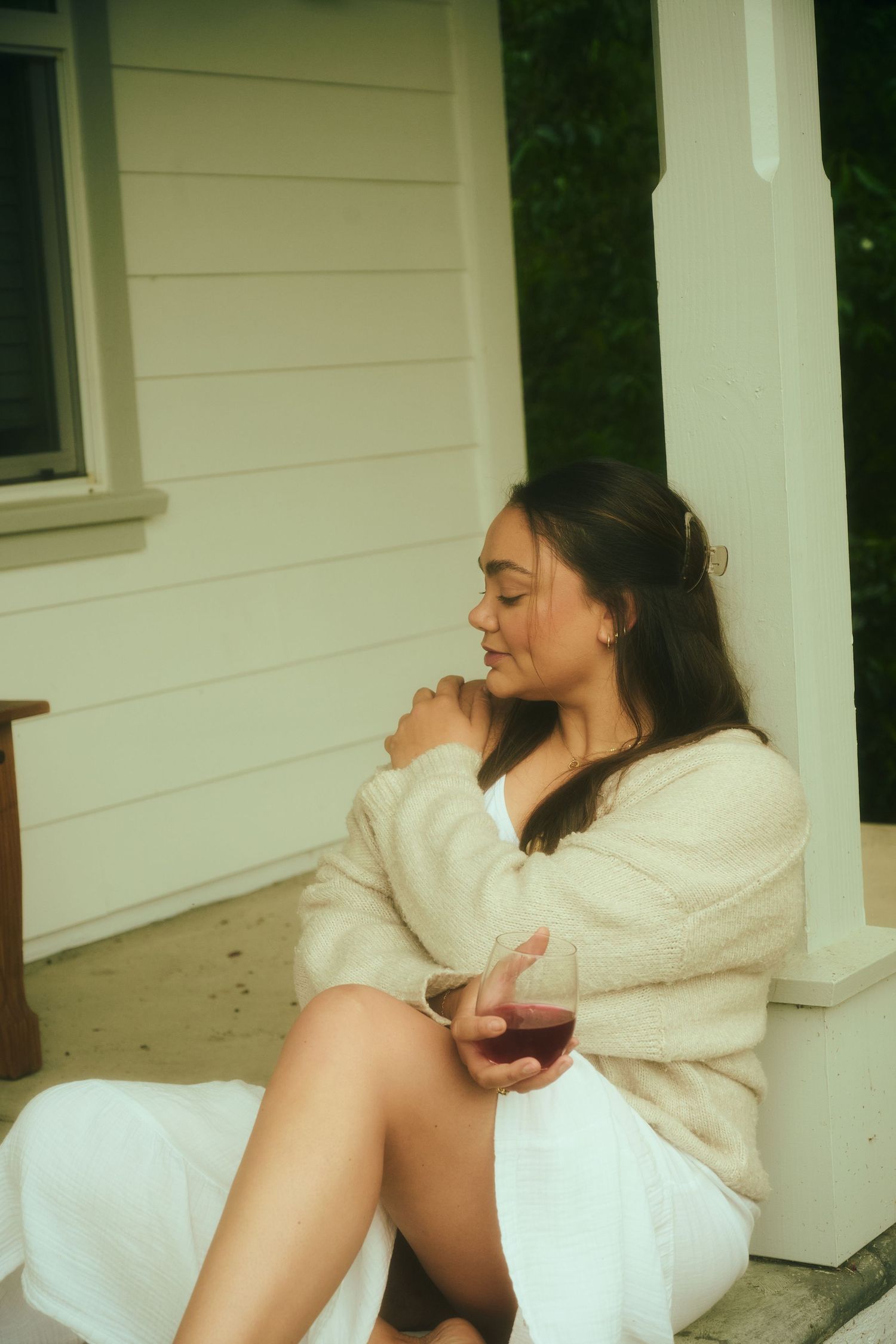 Woman in cream cardigan and white skirt sits on porch steps holding a glass of red wine.