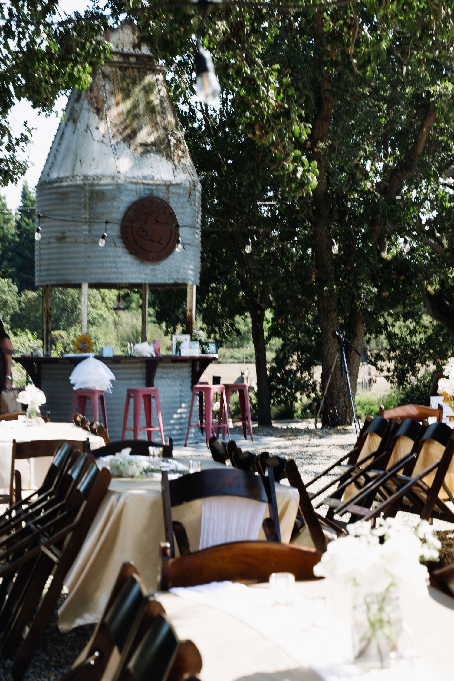 Rustic outdoor wedding venue with wooden chairs, white tablecloths, a vintage silo bar, and red stools under shady trees.