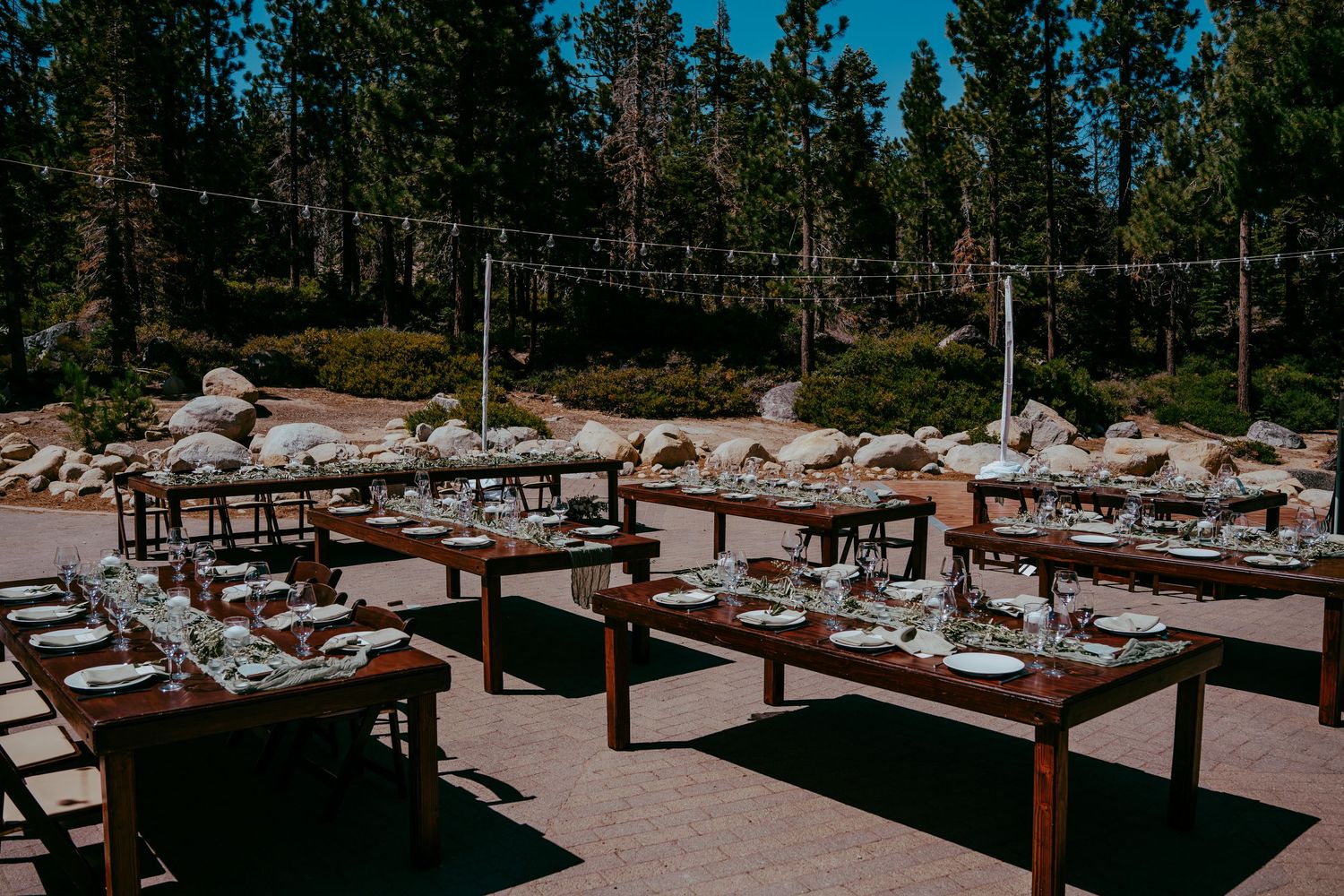 Outdoor rustic wedding reception with wooden farm tables set for dining, surrounded by pine trees and string lights.