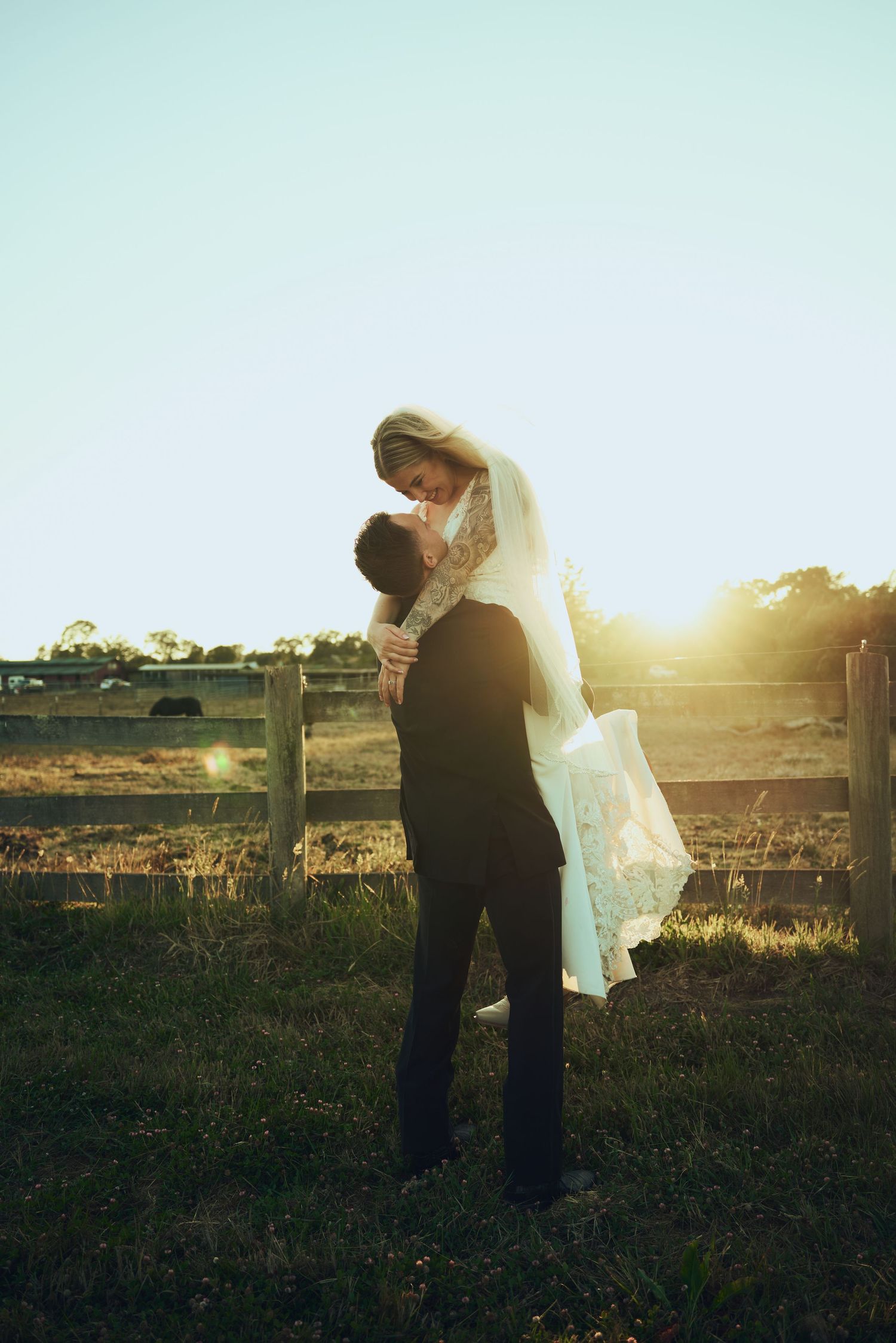 Bride and groom embracing at sunset near a rustic fence, golden backlight illuminating their wedding attire.