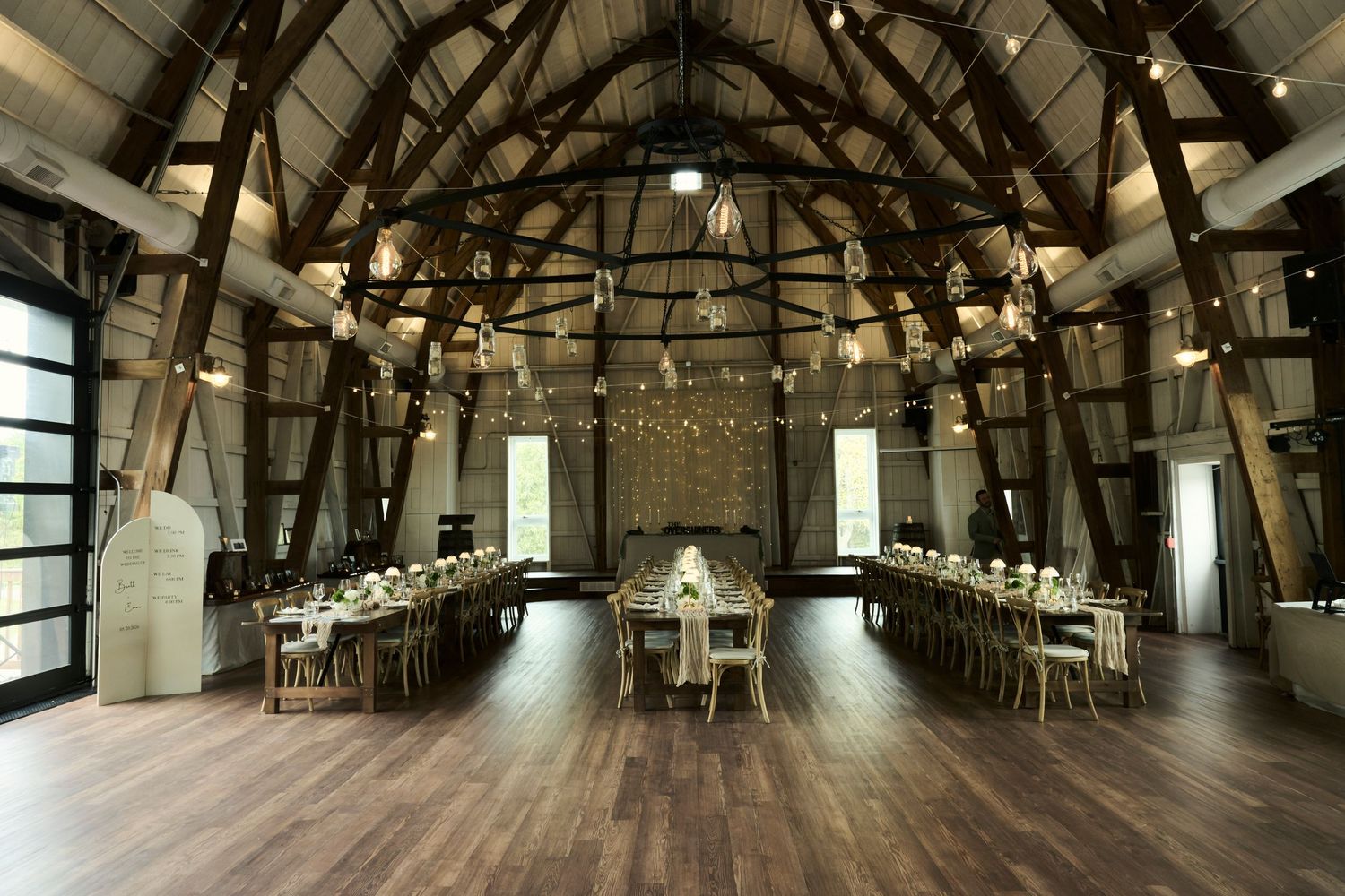 Rustic barn wedding venue with long reception tables, string lights, and wooden beam ceilings on hardwood floors.