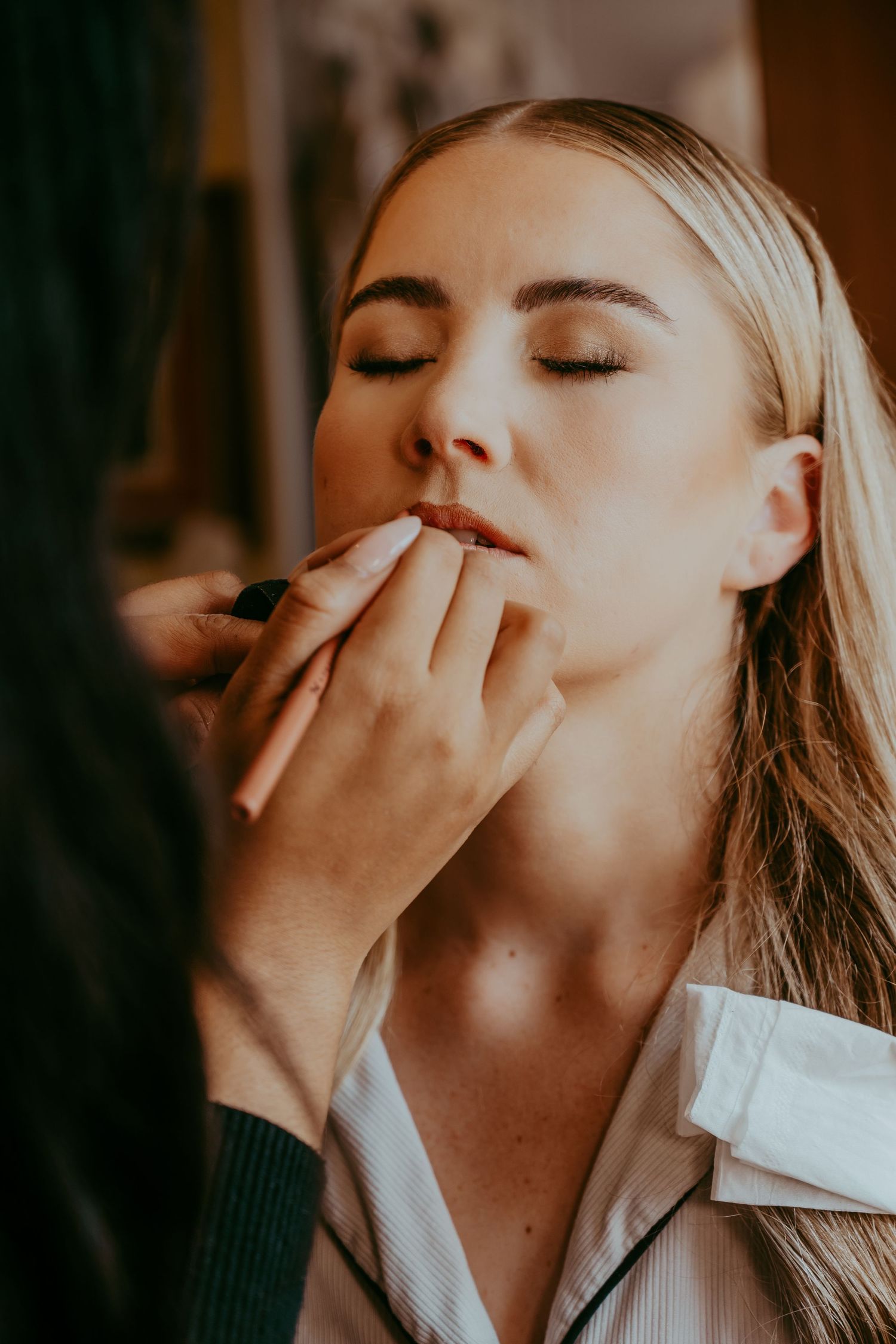 Makeup artist applying lipstick to a young blonde woman with closed eyes during a professional beauty session.
