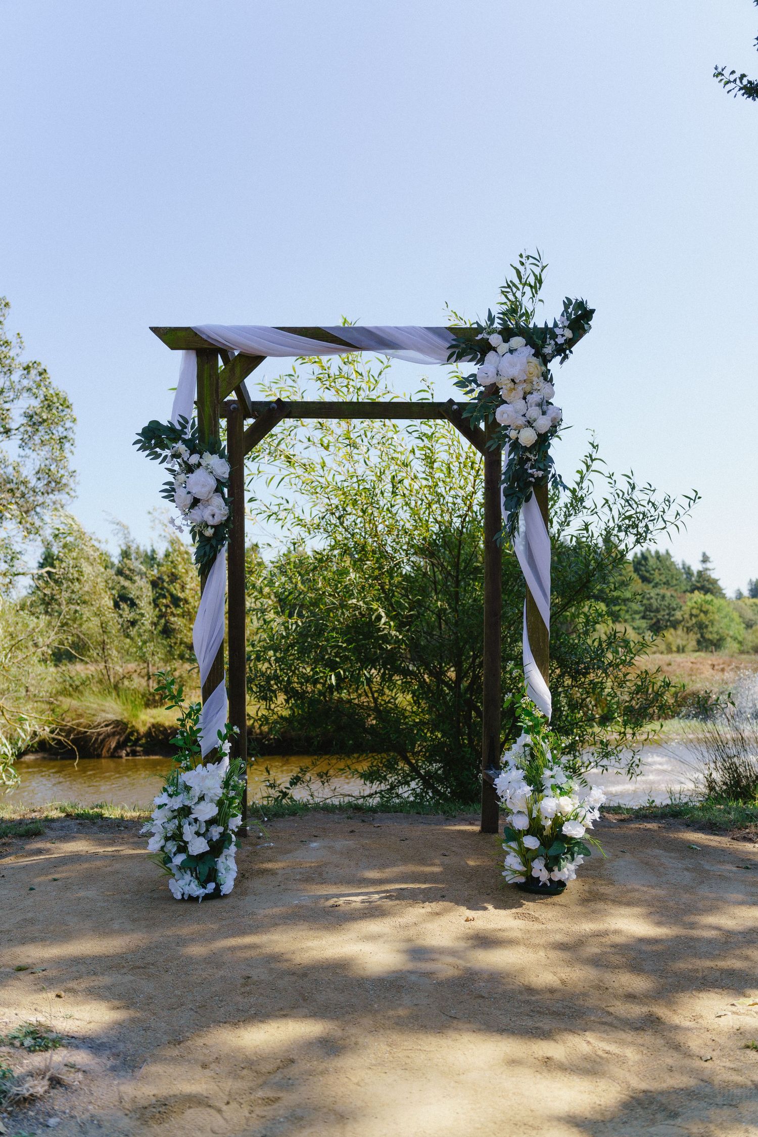 Wooden wedding arch decorated with white flowers and ribbons, set outdoors near a pond surrounded by lush green trees.