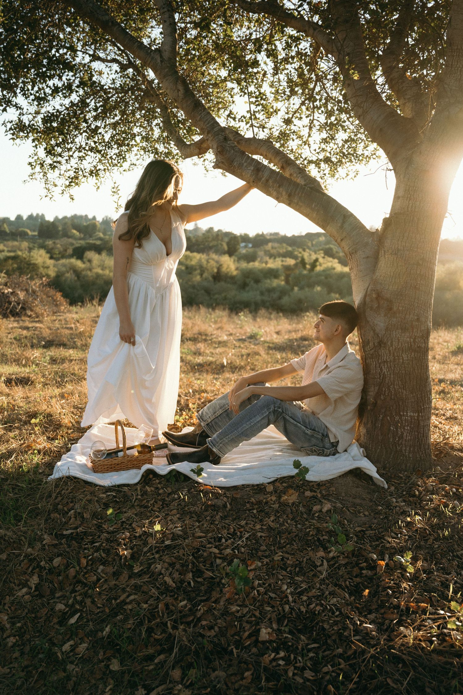 A romantic picnic scene under a tree at sunset with white blankets and natural lighting.